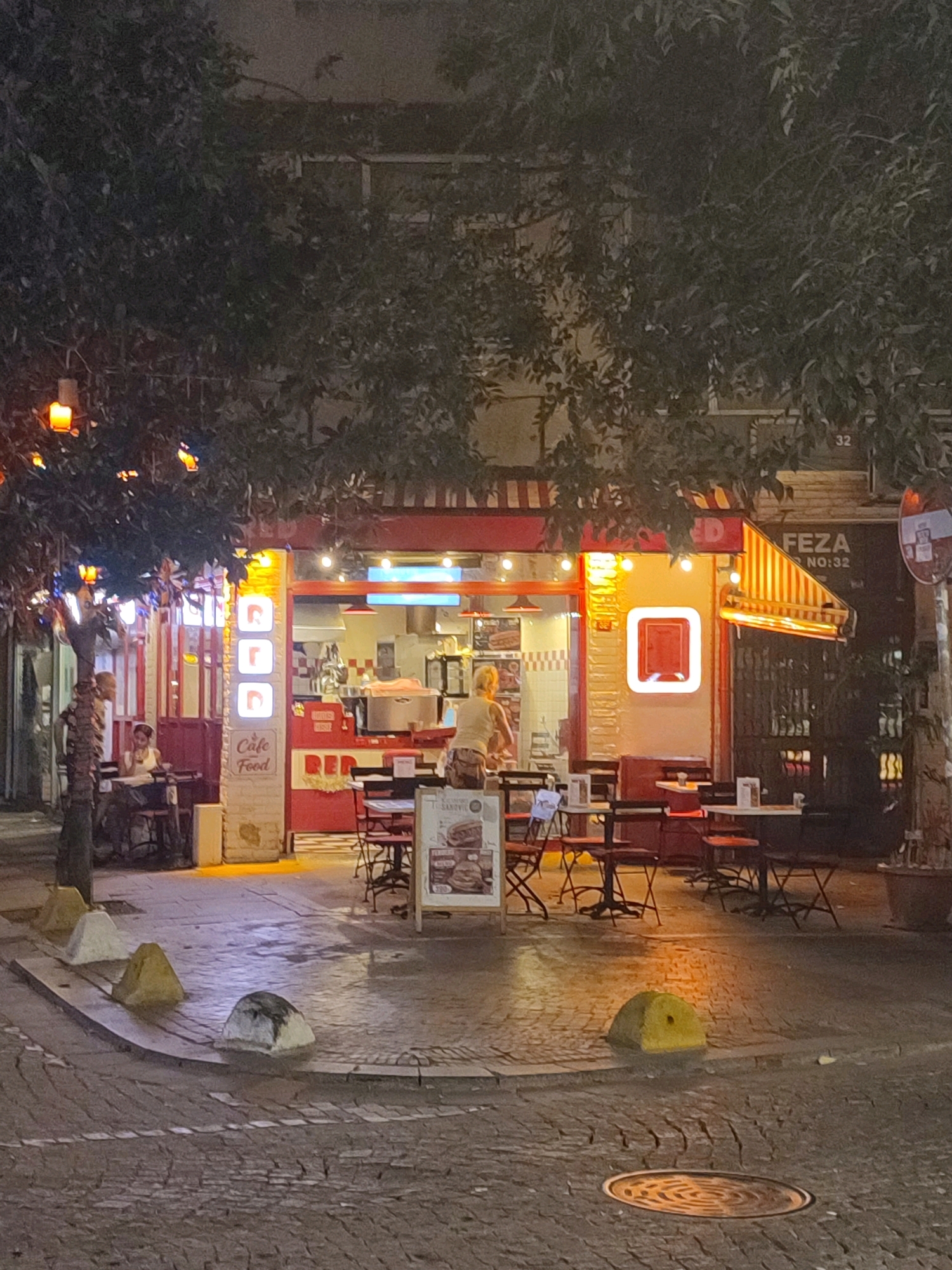 This image depicts a street-level view of a cafe or restaurant exterior at night in Kadıköy, Turkey. The establishment, identified by signage as "Cafe Food RED," features a bright red facade with an awning and outdoor seating. Several tables and chairs are arranged on the cobblestone sidewalk. A person is visible inside the establishment, seemingly working behind the counter, and another person is seated at a table on the left side of the image. Decorative string lights are visible overhead, casting a warm glow. The street is paved with cobblestones, and several concrete bollards are placed along the curb. In the background, building facades are visible, including signage indicating "FEZA NO:32." The scene appears to be on a corner lot, with trees providing some natural canopy.