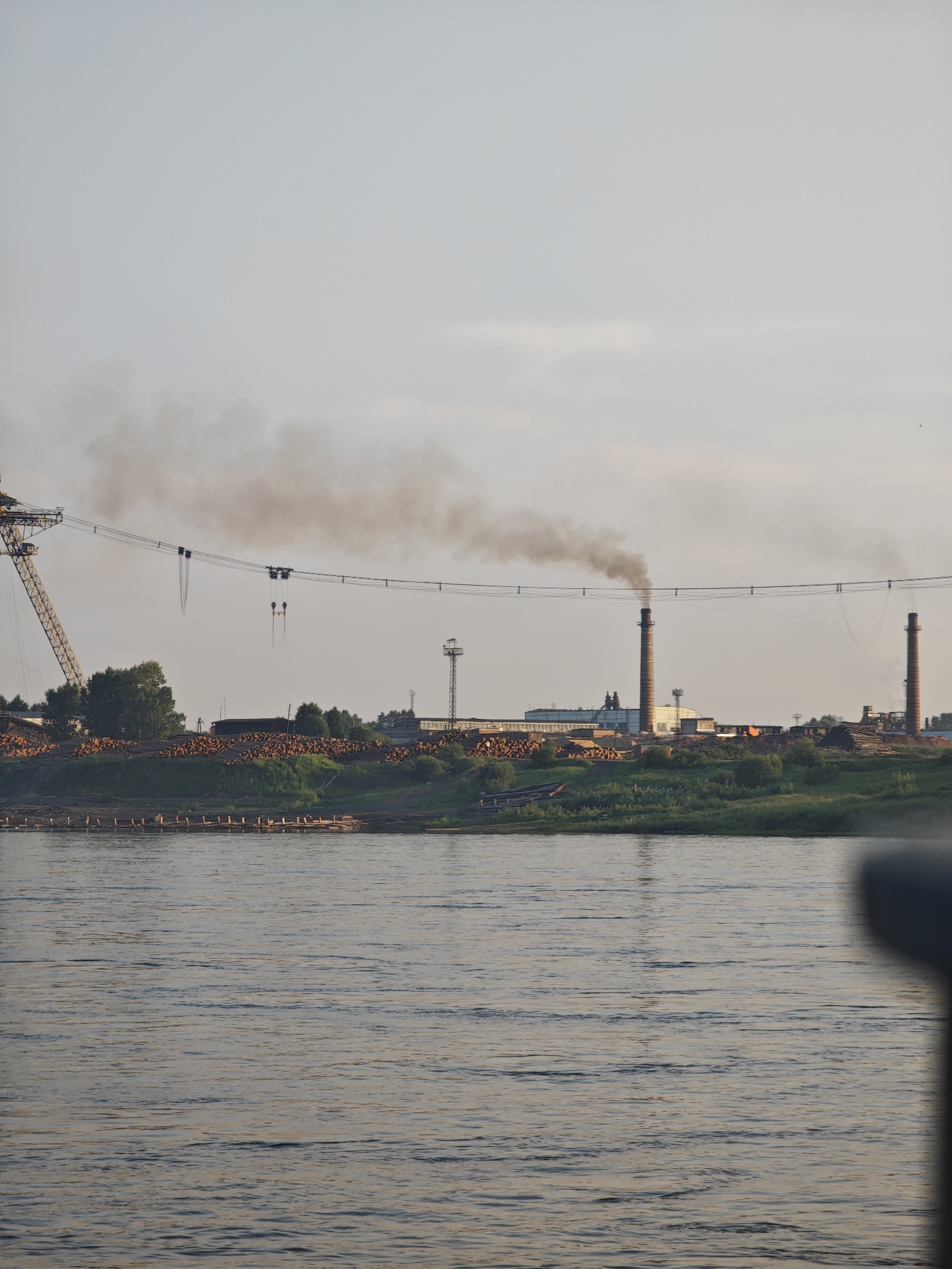 The image captures a scene on the Yenisei River in Lesosibirsk, Russia. The river occupies the foreground, its surface rippled by a gentle current. On the far bank, a sprawling industrial complex is visible. Prominent structures include several tall brick chimneys, one of which is emitting a thick plume of dark smoke into the hazy sky. In the middle ground, a large quantity of logs is stacked along the riverbank, suggesting a lumber or wood processing facility. To the left, a large yellow industrial crane stands, its boom extended over the area. The background is filled with the muted tones of a hazy sky and distant trees. There are no people visible in the image.
