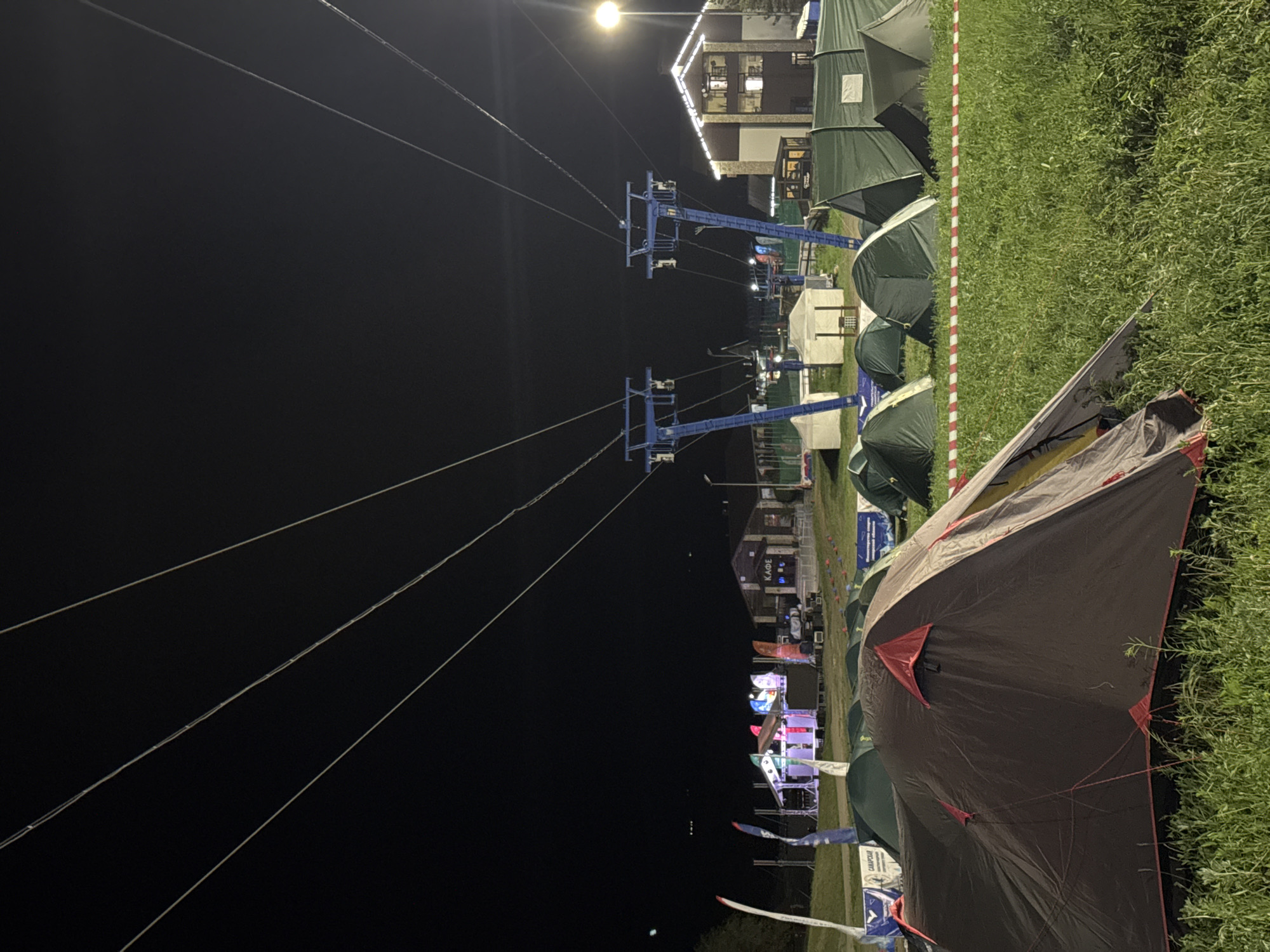 Captured at night, the scene features a campground in Khvalynsk, Russia, situated next to a ski lift. A row of green tents stretches along a grassy area marked with a red and white striped barrier. In the foreground, a large, partially obscured brown tent dominates the view. Beyond the tents, a ski lift with blue support structures is visible, ascending towards a building with illuminated windows. The night sky is dark, with the lift cables cutting across it, illuminated by an overhead light. Several other light sources are visible in the background, adding to the ambience of the scene.