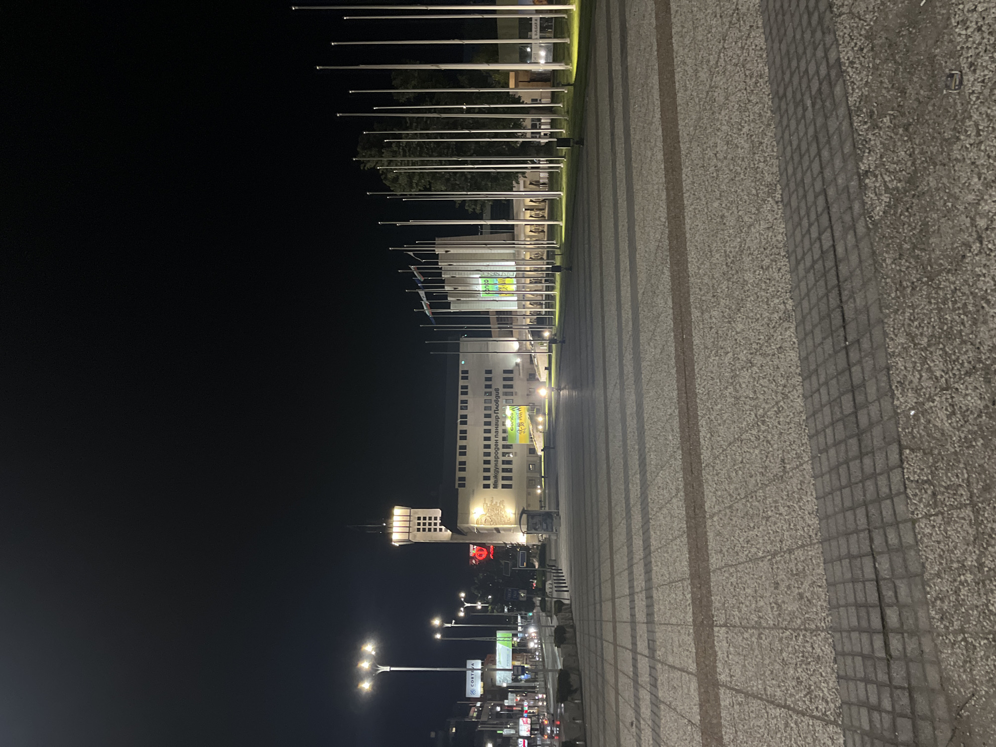 The image shows a nighttime scene in Plovdiv, Bulgaria, featuring a long plaza leading to a building. The plaza is paved with gray stones, arranged in a striped pattern that creates a sense of perspective toward the building. Along the left side of the plaza, a series of flagpoles without flags stretch into the distance. The building is multi-storied with multiple windows and is illuminated with exterior lights. Further down the street, there are streetlights and various signage. The sky is dark.