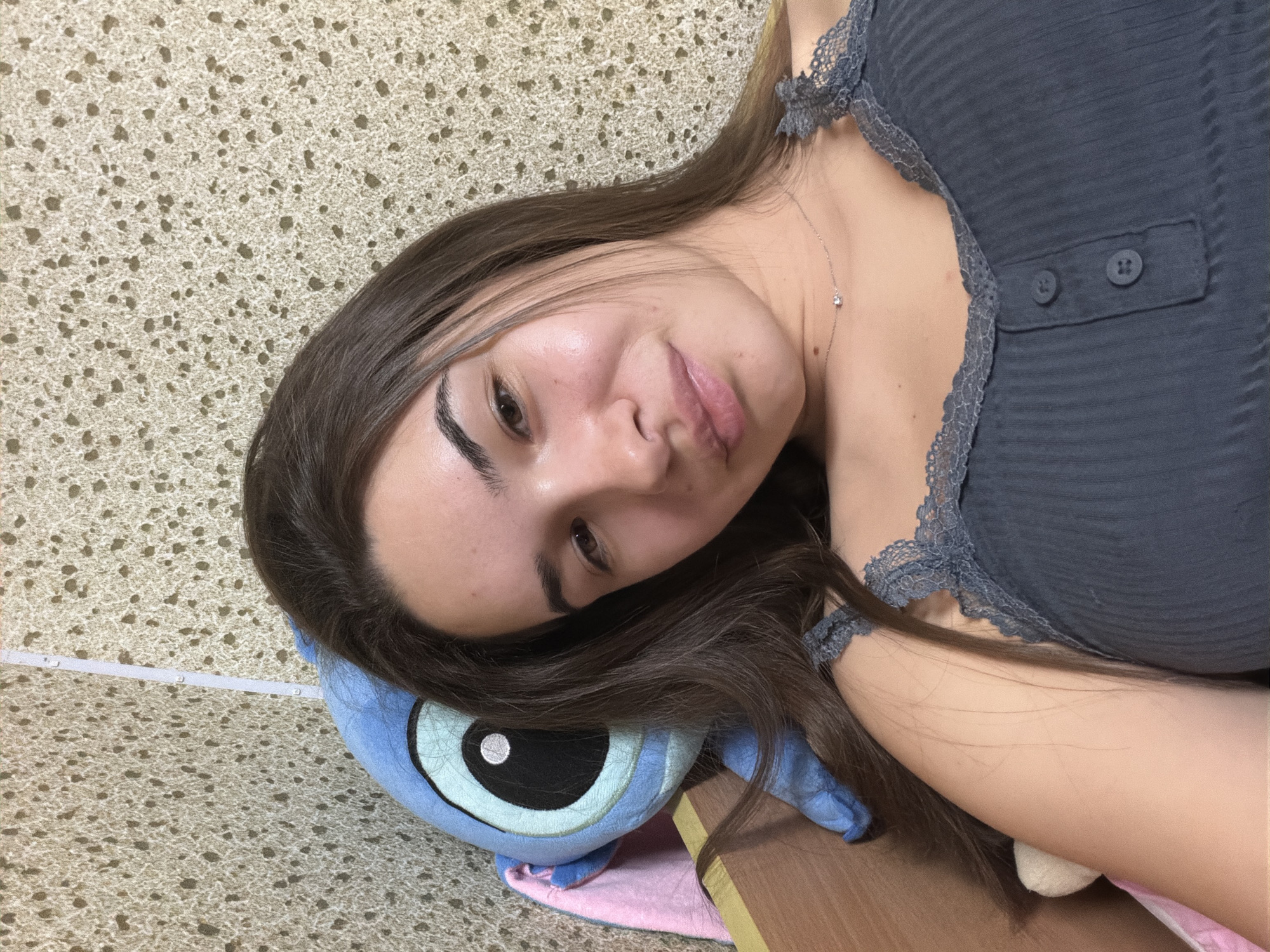 A young woman is lying on her side, her head resting on a blue and pink stuffed animal toy. She is wearing a gray ribbed tank top with lace trim and a delicate necklace. Her long, brown hair is partially covering her face and the toy. The background appears to be a textured beige surface. The scene does not provide enough information to determine a specific location in Sofia, Bulgaria.