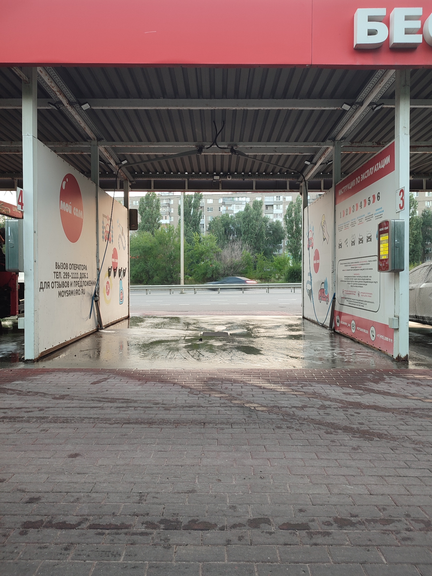The image shows an outdoor car wash area in Voronezh, Russia. The car wash appears to be a self-service station. The foreground is a brick pavement, wet from recent use, with visible tire marks. Two open-air bays are visible, covered by a metal roof. The left bay has a white panel with Russian text and graphics, including a red circle with the word "МОЙ САМ" (Moy Sam - Wash Yourself) and contact information. A long pole with a brush or spray attachment rests against this panel. The right bay features a similar white panel with Russian text, numbered instructions from 1 to 6, and graphics depicting car washing stages. A red box with a yellow display is mounted on this panel, likely a payment or control unit. In the background, trees and apartment buildings are visible, suggesting an urban or suburban setting. A light-colored car is partially visible in the right bay, undergoing or having undergone a wash. The overall environment is overcast.