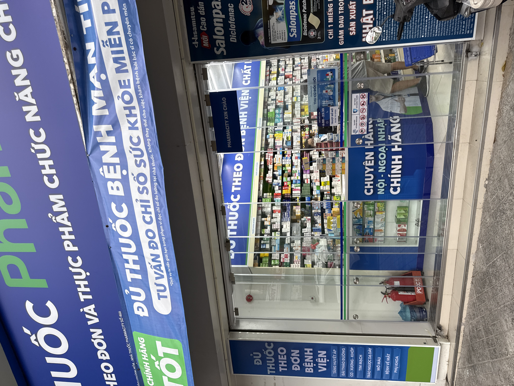 The image shows the exterior of a Pharmacycity store in Nha Trang, Vietnam. The storefront features a large blue and white banner with Vietnamese text, indicating that the store offers a variety of medications and health products. The banner also highlights "CHÍNH HÃNG TỐT" (Genuine and Good) and "ĐỦ THUỐC BỆNH MẠN TÍNH SỨC KHỎE MIỄN PHÍ" (Full of chronic disease medicine for free health).
The entrance to the pharmacy is a glass door with blue signs listing different medical categories like "TĂNG HUYẾT ÁP" (High Blood Pressure), "ĐÁI THÁO ĐƯỜNG" (Diabetes), and "TIM MẠCH" (Cardiovascular). Inside, shelves are stocked with numerous pharmaceutical products.
On the right side of the entrance, another banner advertises "Salonpas" Diclofenac patches. A man wearing a white coat is reflected in the glass door, and a shopping basket is visible inside. A fire extinguisher is placed near the entrance. The overall environment appears to be a typical urban street scene in Vietnam.
