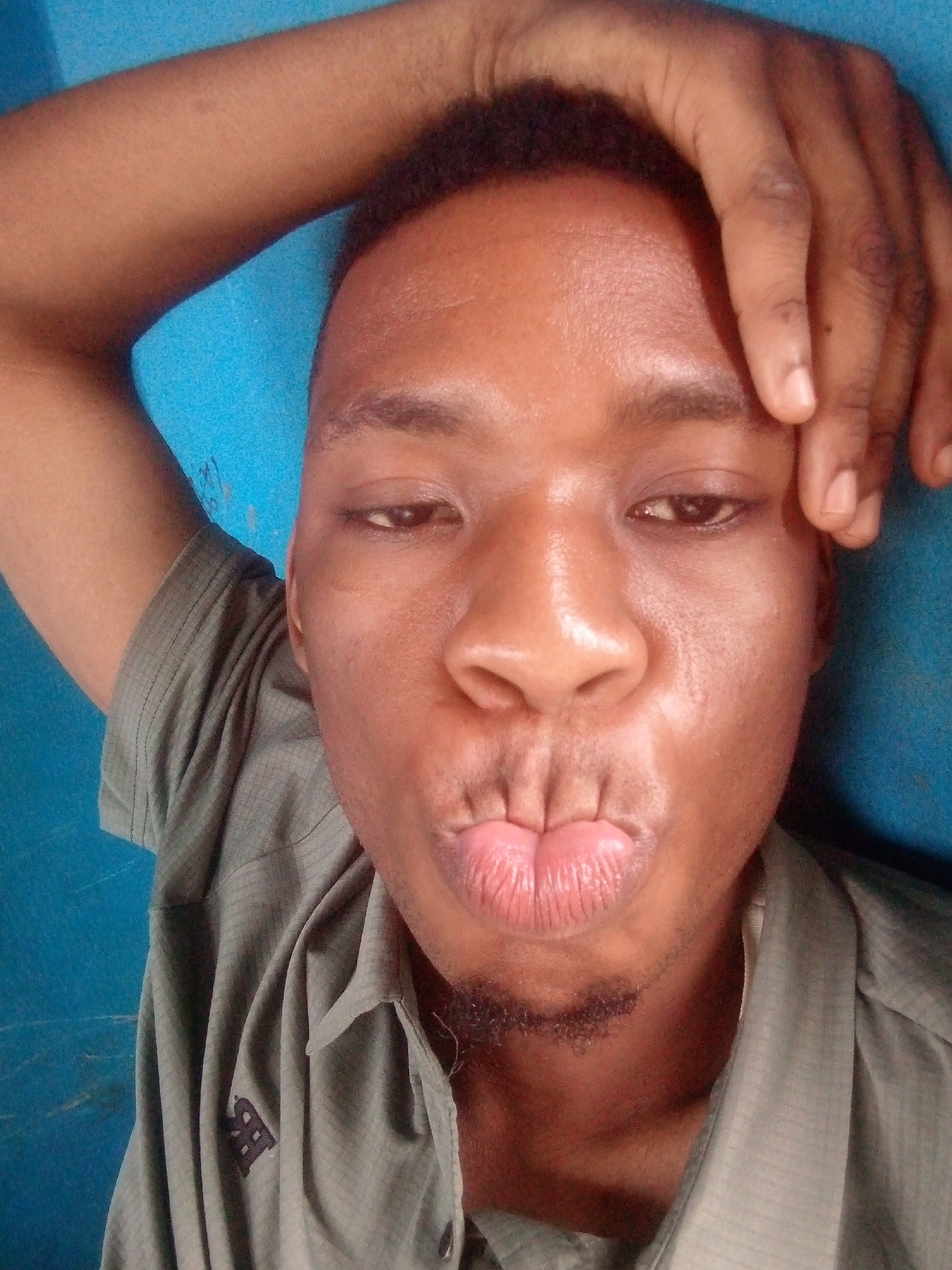 A close-up shot depicts a dark-skinned man with short hair and a light beard, wearing a khaki-colored shirt. He has one arm raised above his head, with his hand touching his forehead. The background is a solid blue wall. His lips are pursed in a kissing position. The location is Unknown, Unknown.