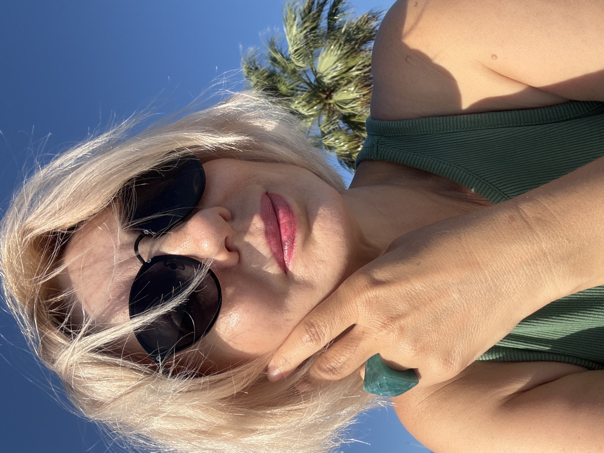 A woman with blonde hair and sunglasses is seen outdoors against a clear blue sky. A palm tree is visible in the background. She is wearing a green top and a large teal ring on her finger. The image appears to be taken on a sunny day. The woman's face is close to the camera, with one hand touching her face. She has pink lipstick on.