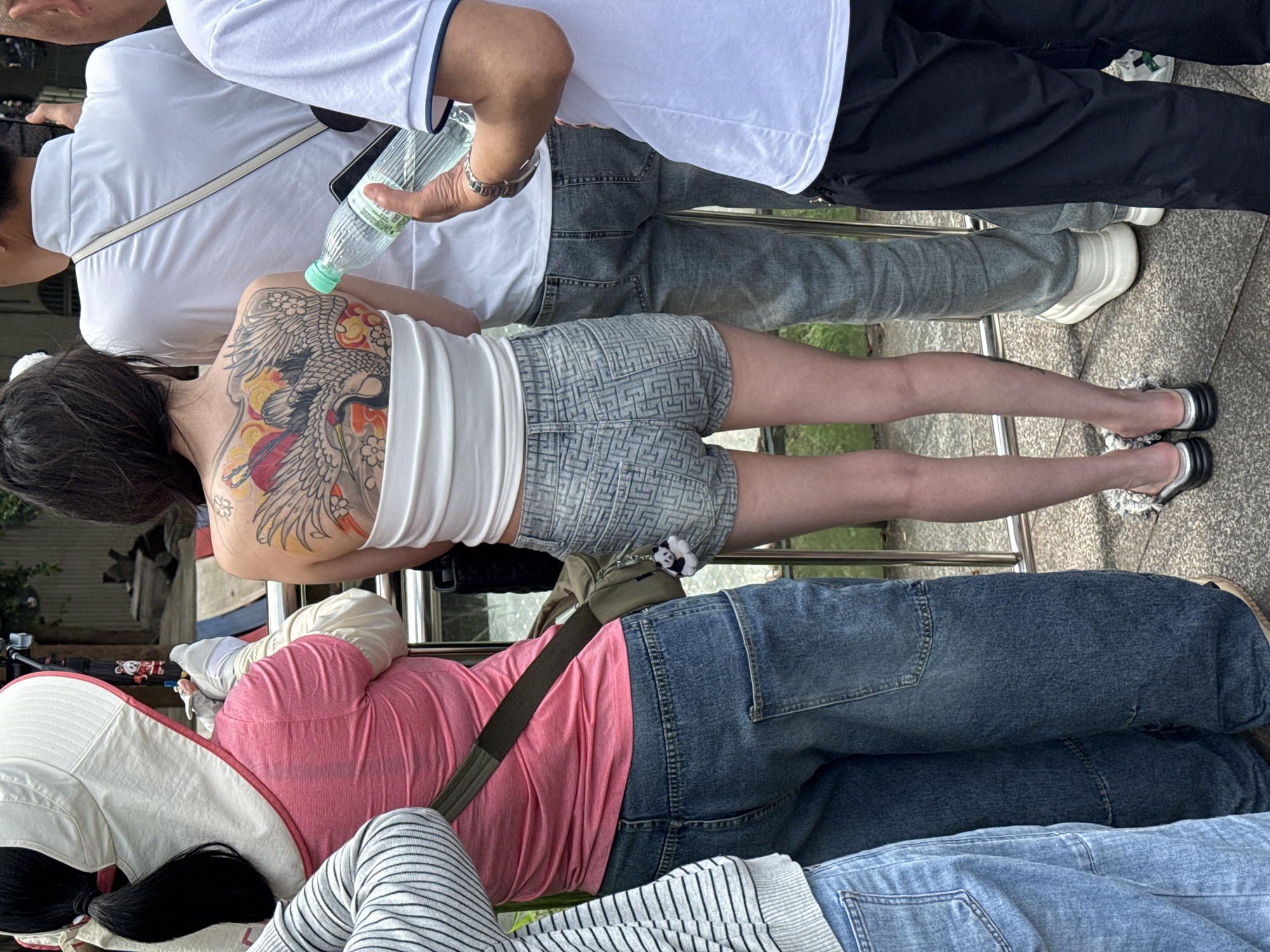 The image shows a group of people standing closely together outdoors. A woman with a large, colorful tattoo on her upper back wears a white tank top and patterned denim shorts and is seen from behind. To her right is a person in a white shirt and light gray denim jeans, holding a water bottle. In front of the woman with the tattoo is a person wearing a pink shirt and blue jeans. The photograph appears to be taken in a public area with a metal railing visible. It is taken in the city of Jiulongpo District, China. The people appear to be observing something.