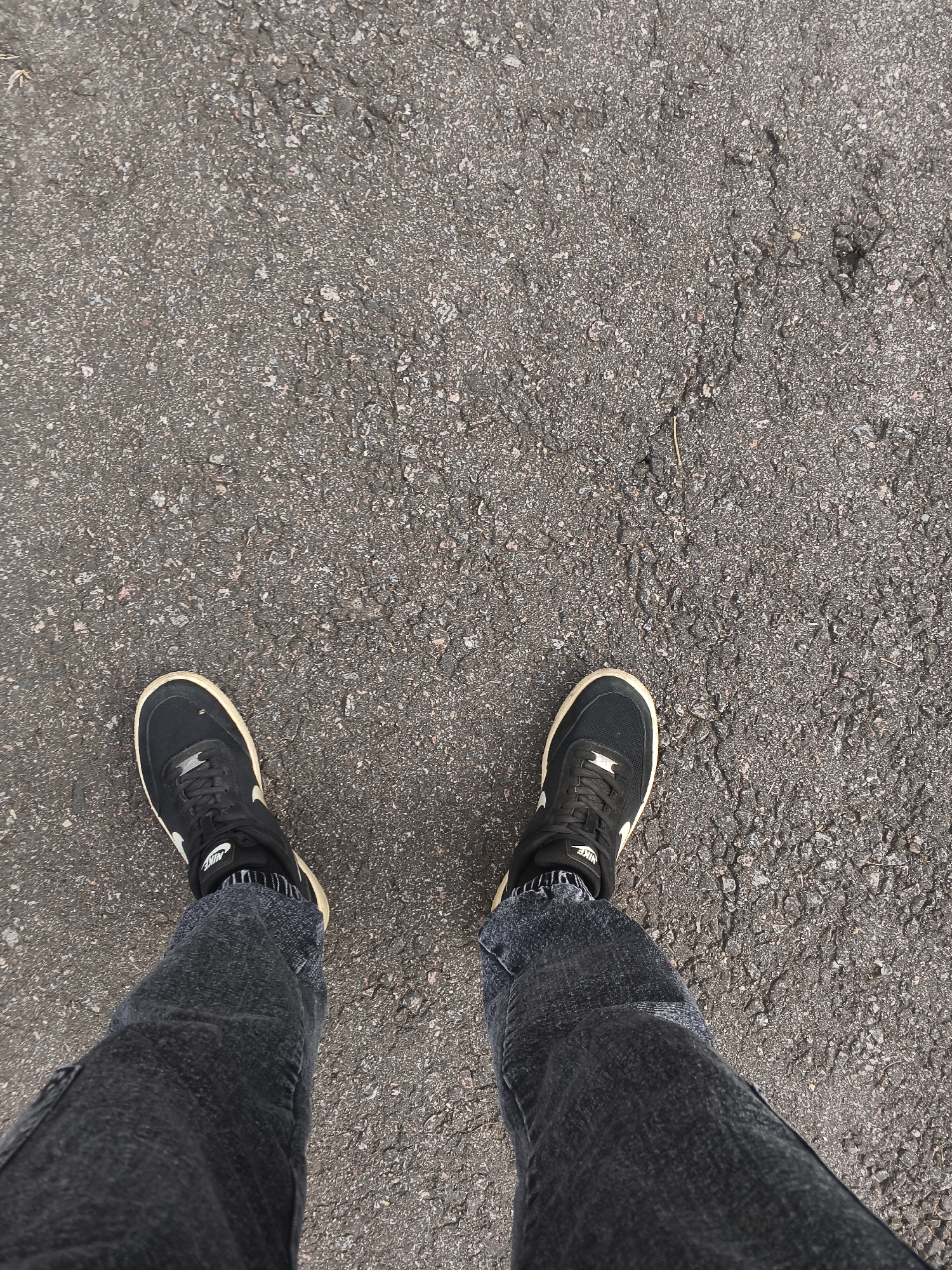 The image shows a pair of feet wearing black Nike sneakers and dark wash jeans, standing on a textured asphalt surface. The perspective is from above, looking down at the person's lower legs and feet. The asphalt appears to be a public walkway or road. There is a small piece of dried grass or straw visible on the asphalt in the upper right portion of the image. No other people, animals, or significant objects are visible. The lighting suggests it is daytime. The location is identified as Mokshan, Russia.