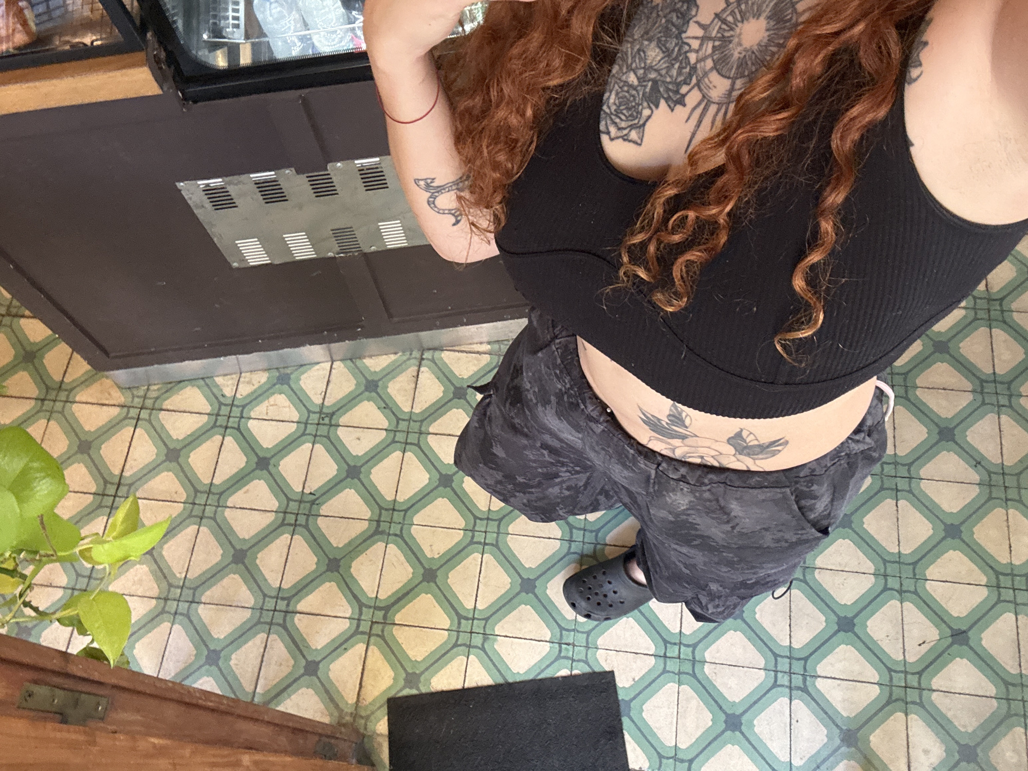 An overhead, high-angle shot captures a person standing indoors. The subject, with long, wavy red hair, wears a black ribbed crop top, dark grey or black cargo-style shorts, and black Crocs. Several tattoos are visible: a large, intricate floral or mandala-like design on the upper chest and left shoulder, a smaller abstract design on the left bicep, and a floral pattern on the lower back, just above the shorts.
The person stands on a patterned tiled floor, featuring light cream squares interconnected by a grid of green and blue-grey lines, forming a repeating diamond motif. To the left, a vibrant green leafy plant is partially visible. In the upper part of the frame, a dark counter or cabinet surface is seen, featuring a rectangular metal grate. Above this, a reflection in a glass surface suggests a display case or appliance. A small black doormat is on the floor in the lower-middle, with a wooden frame visible at the very bottom left.
The setting appears to be an interior space, likely a cafe or shop entrance, possibly in Buenos Aires, Argentina, consistent with the characteristic tile work. The lighting is bright and even, suggesting daytime. The person is simply standing; no specific activity is depicted. No text is explicitly visible in the image.