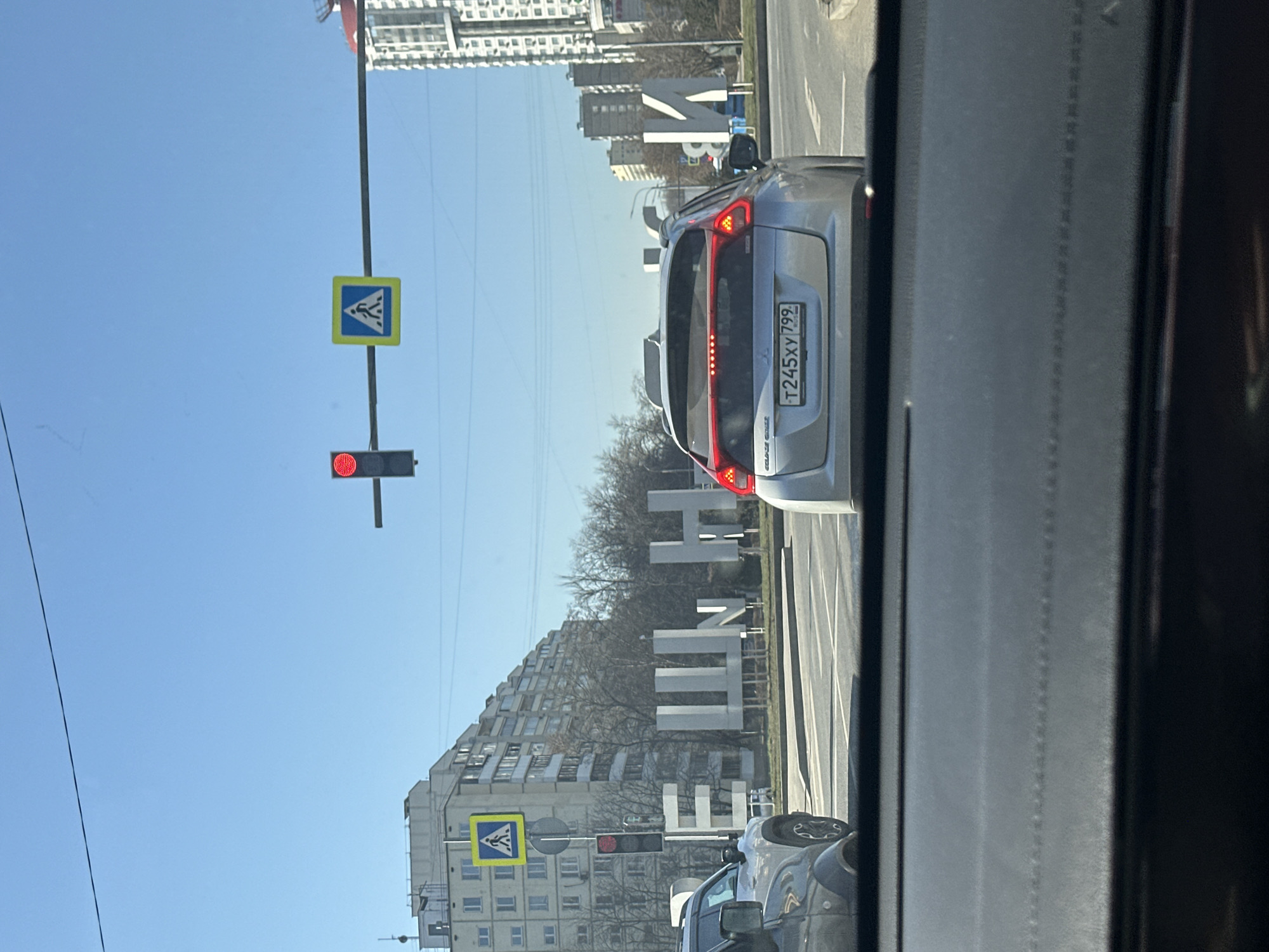 The image, taken from inside a car, shows a street scene in Moscow, Russia. The car is stopped at a red traffic light. The license plate of the car in front reads "T245XY799." The photo captures the "Leninsky Prospekt" monument, a large architectural display spelling out "ЛЕНИНСКИЙ ПРОСПЕКТ" in Cyrillic (Leninsky Prospekt in English). There are buildings on both sides of the street. Pedestrian crosswalk signs are visible, and the sky is clear and blue.