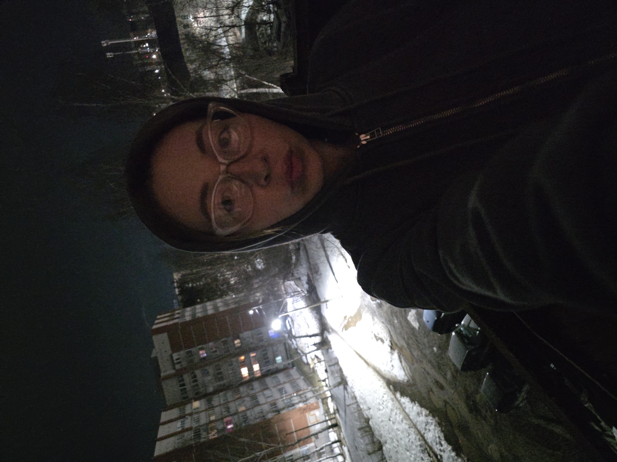 A nighttime selfie of a person with glasses and a hood, likely in Chaikovsky, Russia. The person is positioned in the foreground, and behind them, there's a view of a multi-story building with illuminated windows and streetlights. A snowy yard or street is also visible. In the far background, lights appear to be reflected in water. The image has low lighting and slight motion blur, indicating the photo may have been taken quickly.