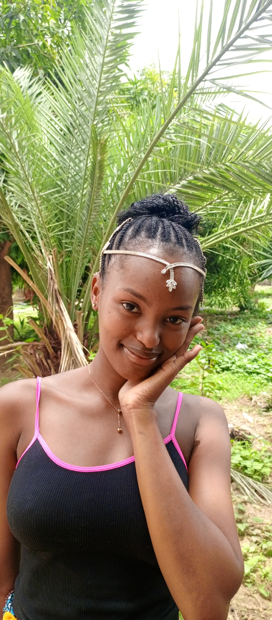 A young woman with dark skin and braided hair poses for a photo outdoors. She wears a black tank top with pink trim, a delicate gold necklace, and a decorative headpiece. Her hand is gently touching her cheek as she smiles. Behind her is a lush, green palm tree and other foliage. The setting appears to be a natural environment, likely a garden or park in Keffi, Nigeria.