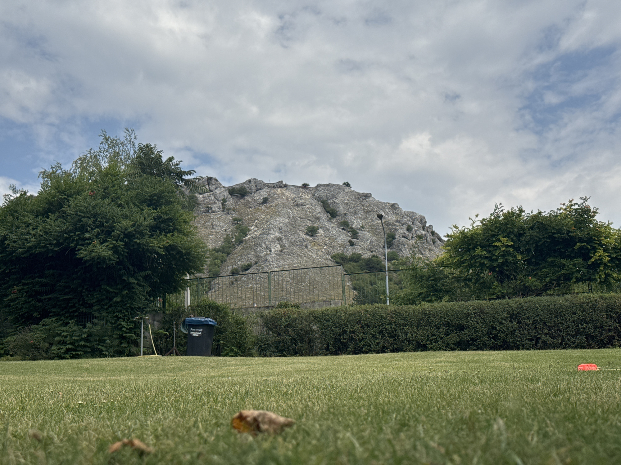 The image depicts a grassy park area in Hainburg an der Donau, Austria, with a rocky mountain in the background. In the foreground, a well-maintained green lawn occupies the majority of the frame. Towards the middle ground, there's a dense hedge, and beyond that, a metal fence runs horizontally across the scene. A black trash bin with "Restmüll" written on it is positioned to the left of the fence, next to a spigot and a yellow hose. A street lamp is visible to the right of the bin. The mountain, a prominent feature, is grey and rugged with sparse vegetation. The sky is overcast with patches of blue sky visible. The overall impression is a serene outdoor landscape, likely a public park or recreational area in Hainburg. There are no people or animals visible in the image.
