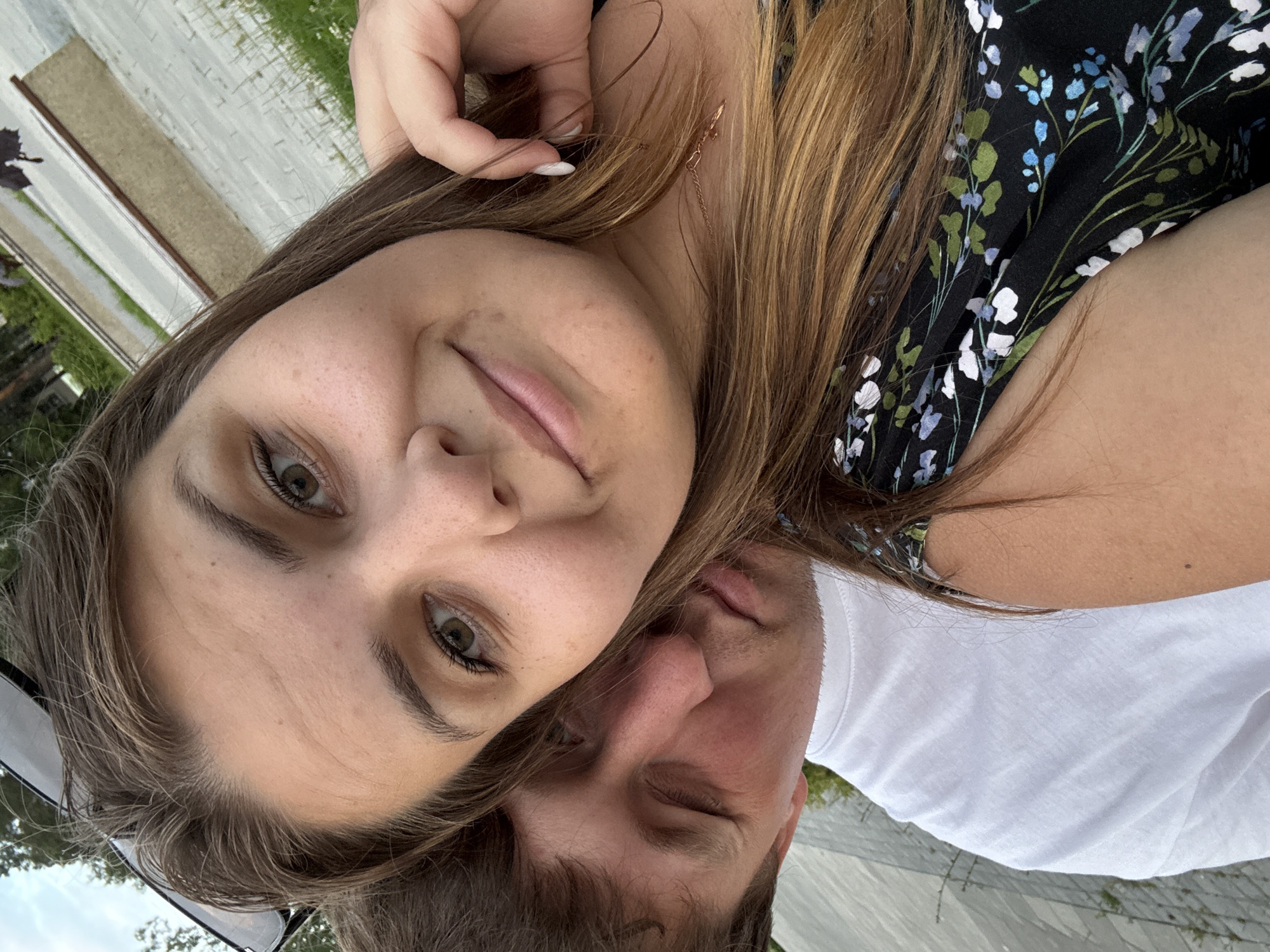 A selfie taken outdoors shows a young Caucasian couple. The woman is in the foreground, facing the camera and smiling. She has long, brown hair, and is wearing a black dress with a floral pattern. The man is positioned behind her, resting his head on her shoulder, with his eyes closed and a slight smile. He is wearing a white t-shirt. The background appears to be an urban park or public space, with paved walkways, grass, and trees visible. A section of what looks like a skateboarding or rollerblading ramp is present in the mid-ground. The lighting suggests it is daytime. The setting is consistent with a park in the city of Chaikovsky, Russia.