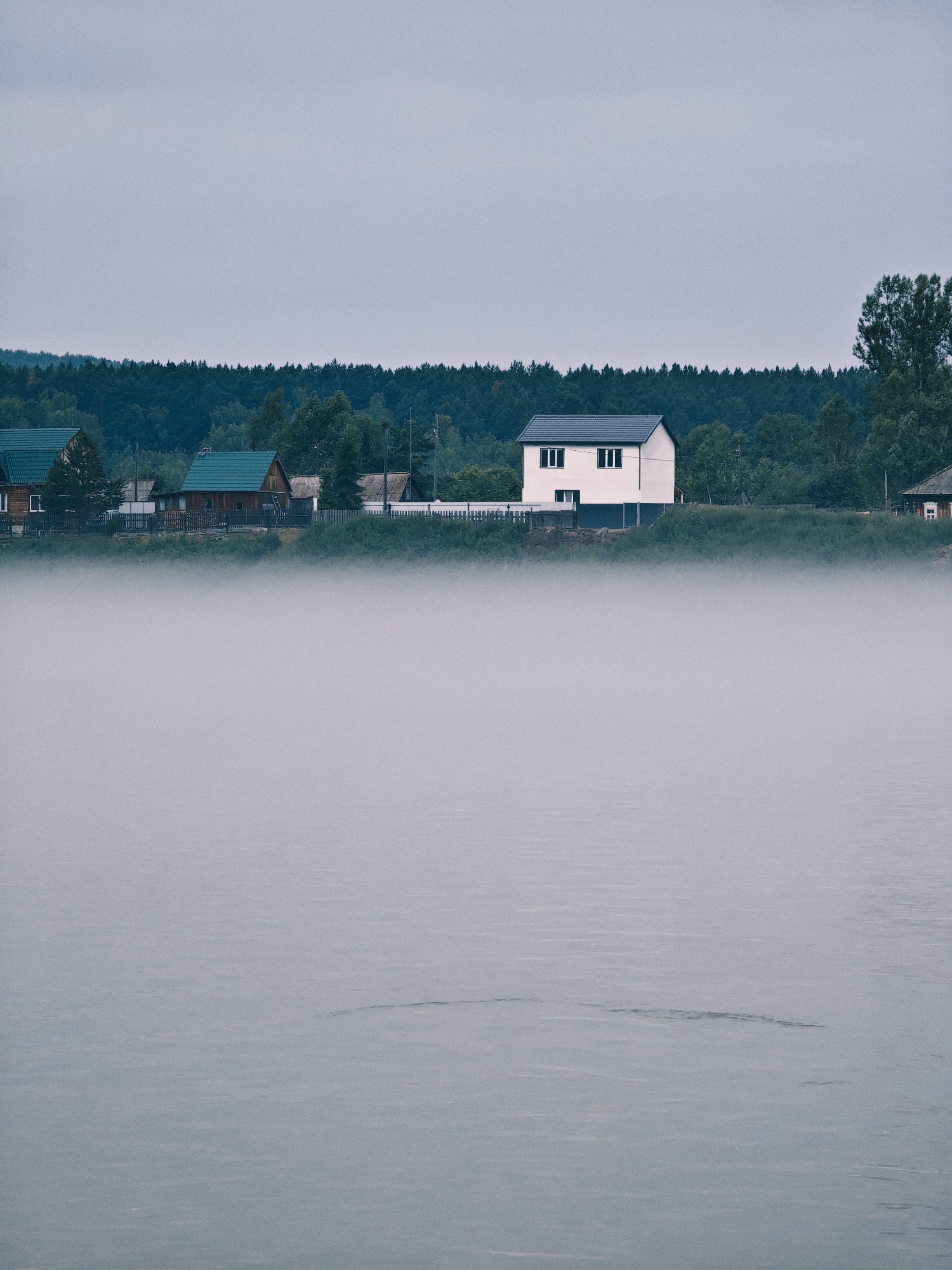 This image depicts a serene scene in the city of Atamanovo, Russia. In the foreground, a body of water is shrouded in a dense layer of fog, its surface disturbed by subtle ripples. Beyond the fog, the opposite bank of the water features a line of houses nestled against a backdrop of dense green forest. Several houses with dark roofs are visible, including a prominent two-story white house with dark-trimmed windows. A wooden fence runs along the bank in front of some of the houses. The sky above is overcast and a pale gray, contributing to the subdued and atmospheric mood of the scene. The overall impression is one of quiet and natural beauty.