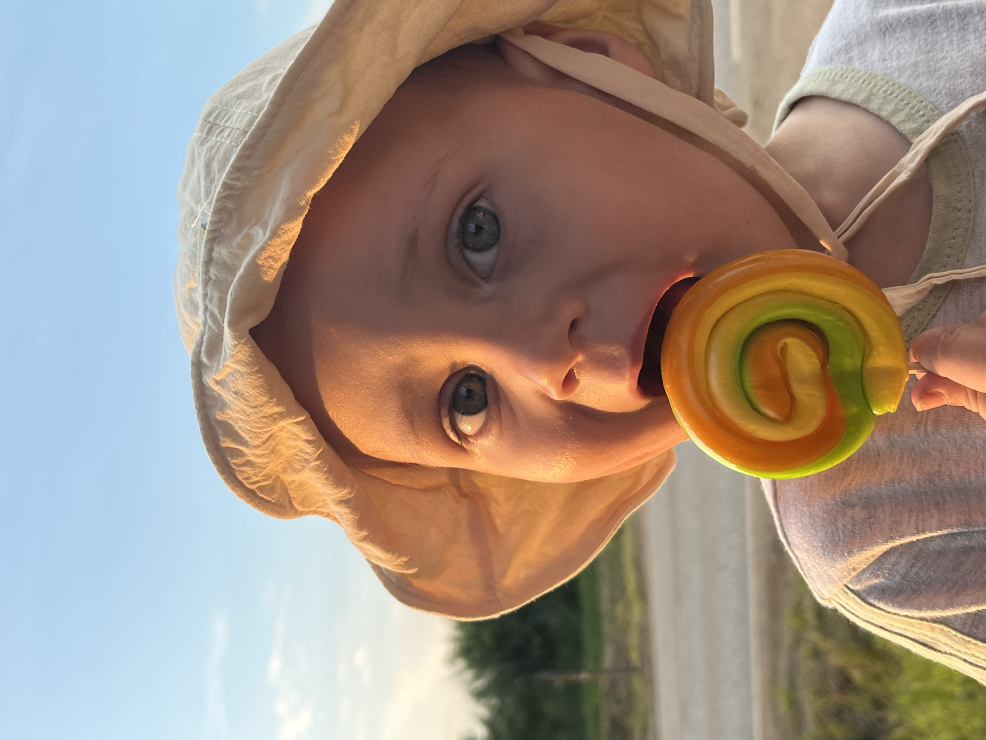 The image shows a close-up of a young child wearing a light-colored sun hat, with a colorful lollipop in their mouth. The child has wide, greenish-blue eyes and is looking towards the camera. The setting appears to be outdoors, with a hazy blue sky and some greenery and a road visible in the background. The sun is setting, casting a warm, golden light on the child's face and hat. There is no indication of the location being in the city of Фоки, Russia, or any other specific location.