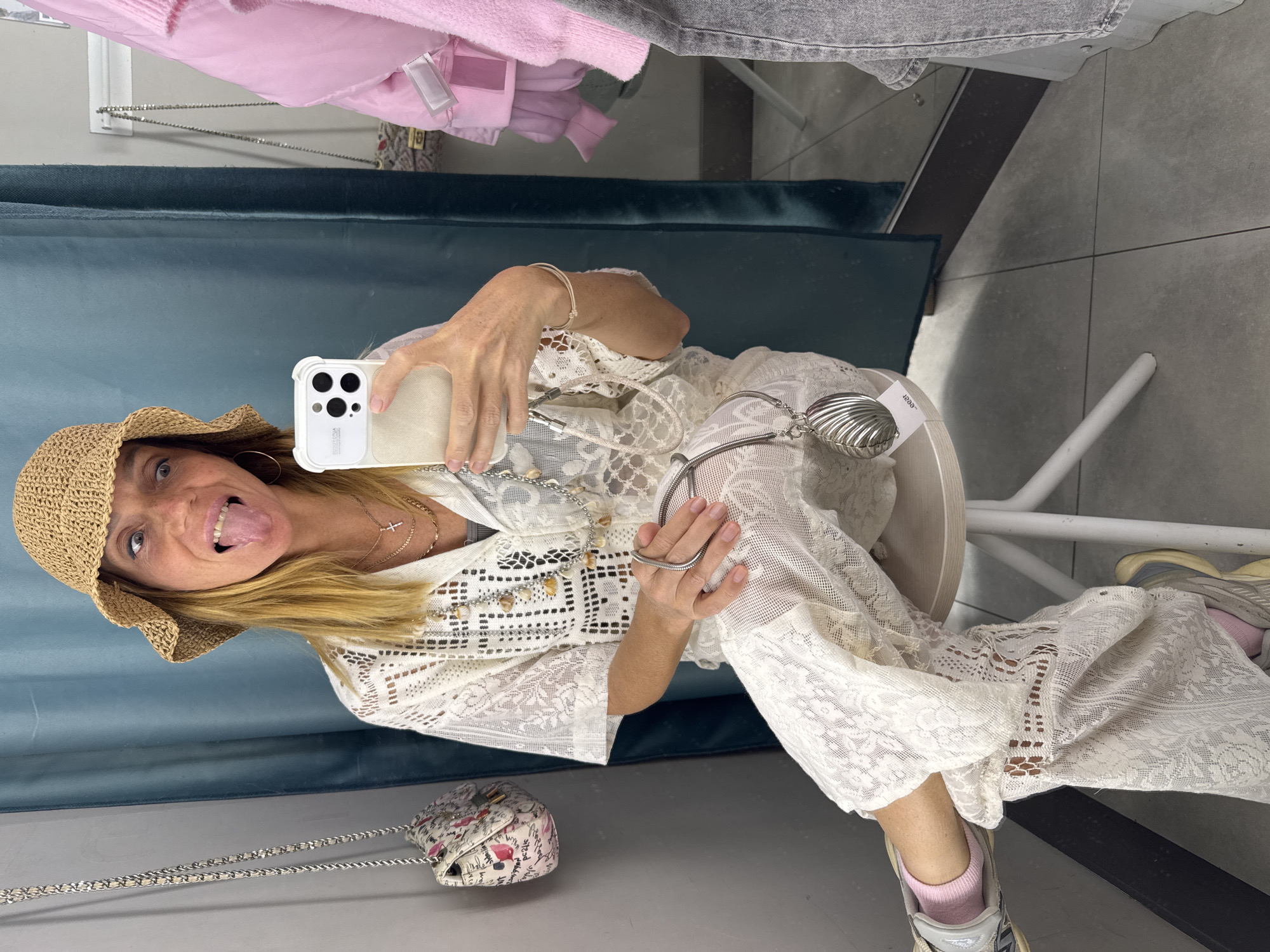 A fair-skinned woman, likely in her 40s or 50s, is taking a selfie inside what appears to be a fitting room. She is wearing a straw hat, a white lace dress or cover-up, and chunky sneakers with pink socks. She is sticking her tongue out, indicating a playful mood. She is holding a cell phone in her right hand, taking the picture. She is seated on a white stool and there is a small floral purse hanging to her left. There is a blue-green curtain behind her, and other clothing items hanging in the periphery, suggesting this is a retail establishment. The tiled floor and modern aesthetic suggest a contemporary store. There is no indication of this image being in the city of Saint Petersburg, Russia.