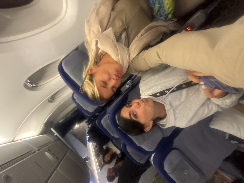 Two women are seated in adjacent airplane seats, posing for a selfie. The woman on the left has blonde hair and is wearing a tan jacket and scarf. The woman on the right has dark hair and is wearing a gray hooded sweatshirt. Both women are making kissing faces. The plane cabin is visible in the background, with other passengers and rows of seats. The image is slightly tilted.