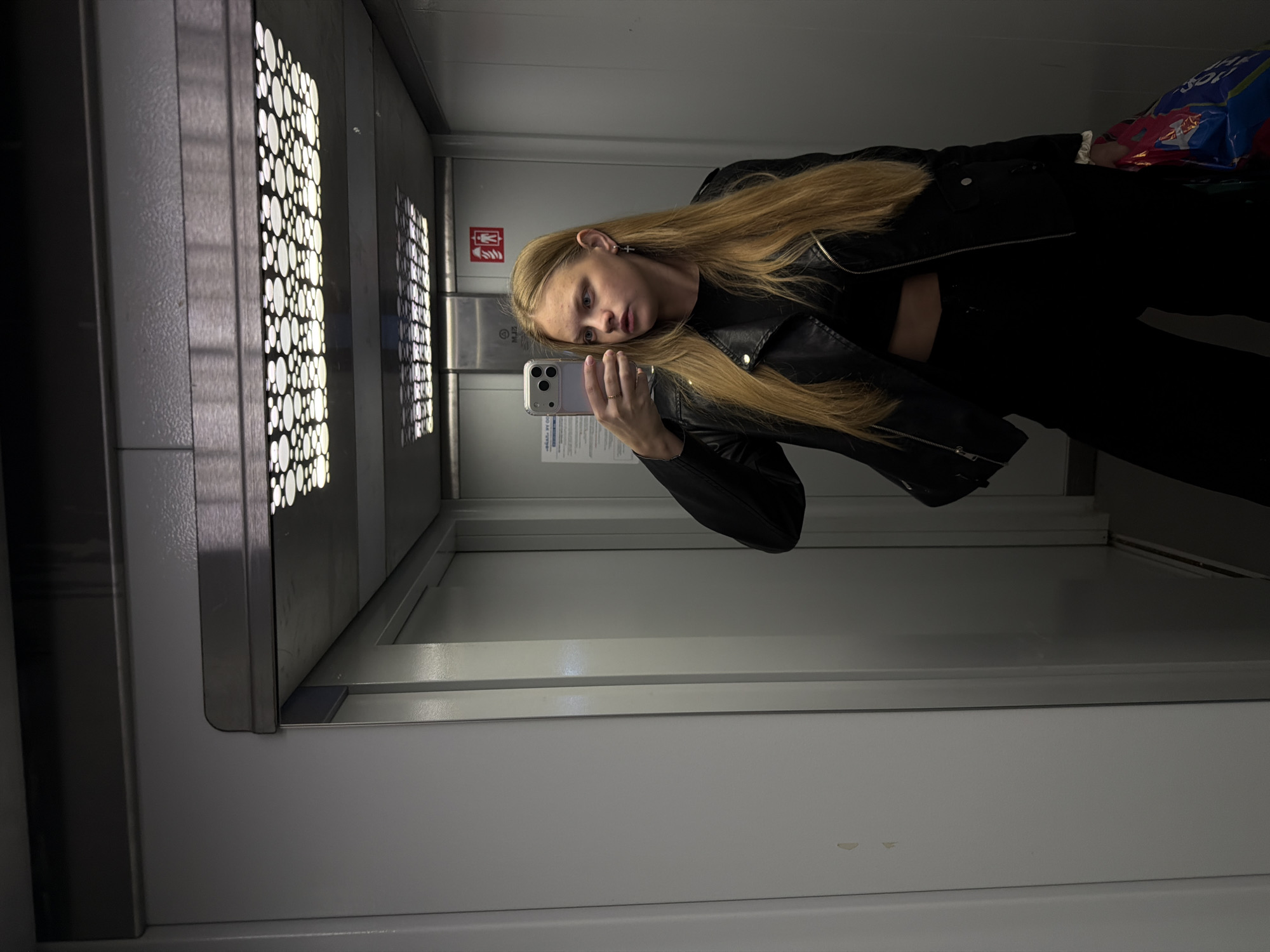 A young woman with long, straight blonde hair is taking a mirror selfie inside an elevator in Izhevsk, Russia. She is wearing a black leather jacket with prominent zippers over a black top that reveals a hint of her midriff, and black pants. She holds a silver smartphone with a clear case in her right hand, aiming it towards her reflection to capture the image. A simple ring is visible on her right hand, and a small cross earring hangs from her right ear. Her expression is neutral, with her gaze directed towards the phone. In her left arm, she carries a colorful plastic bag, predominantly blue and red, with partially visible text that appears to read "SOU" or "SHINE".
The elevator interior features light grey, metallic walls and a reflective floor. The ceiling is equipped with unique rectangular light fixtures, perforated with numerous circular cutouts that emit bright light, casting distinct dotted patterns onto the walls. A red emergency sign, featuring white pictograms resembling fire safety symbols, is mounted on the back wall of the elevator. Faint alphanumeric characters, such as "M22", are also visible on the metallic panel. The overall environment is brightly lit.