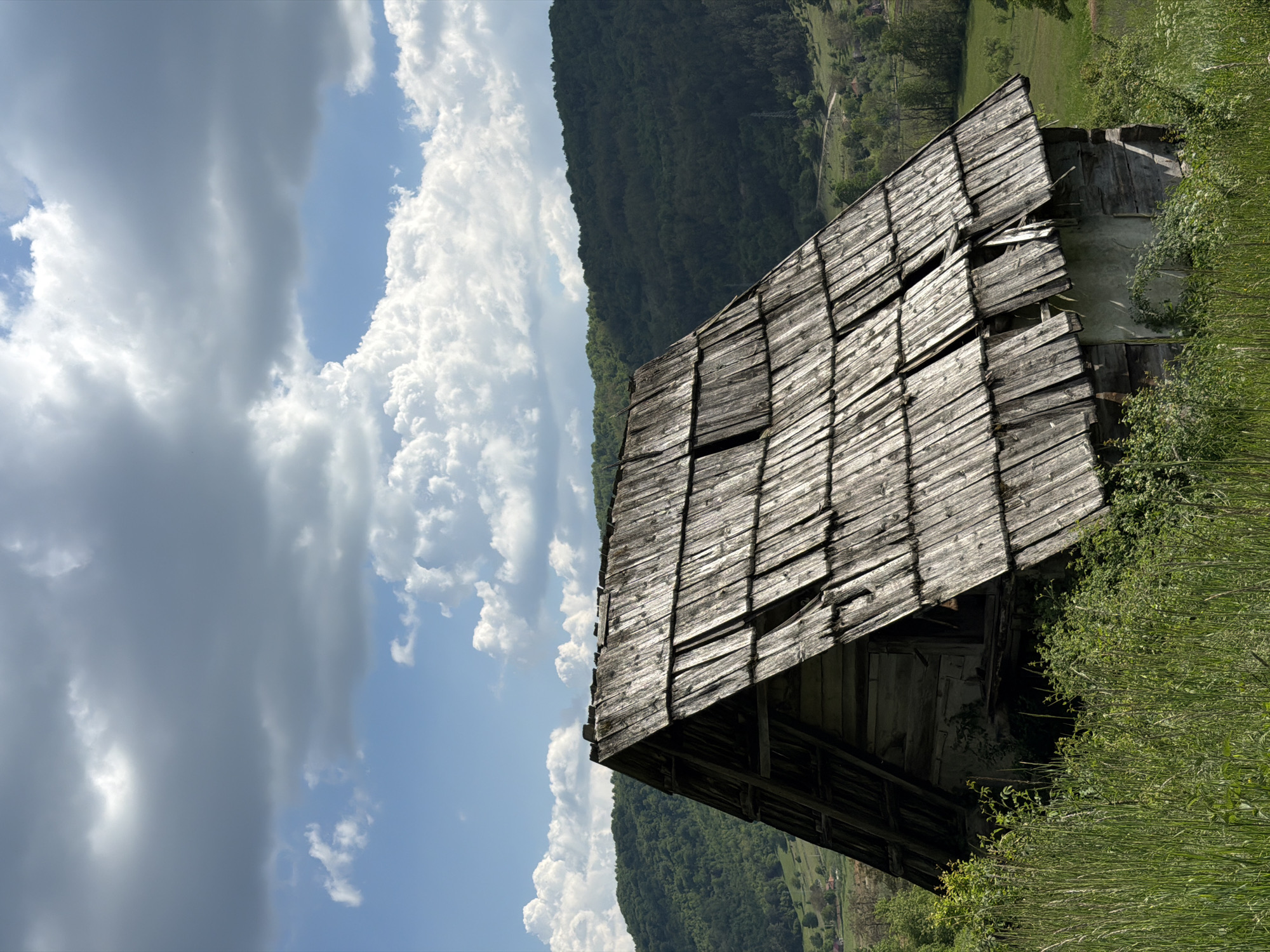 The image shows an old, dilapidated wooden structure, possibly a barn or shed, in Otilovići, Montenegro. The roof is made of weathered wooden planks, with several missing or broken, revealing gaps. The structure is surrounded by tall green grass and vegetation. In the background, there is a lush green mountain landscape under a sky with a mix of blue sky and fluffy white clouds. The overall impression is one of rural decay in a mountainous environment.