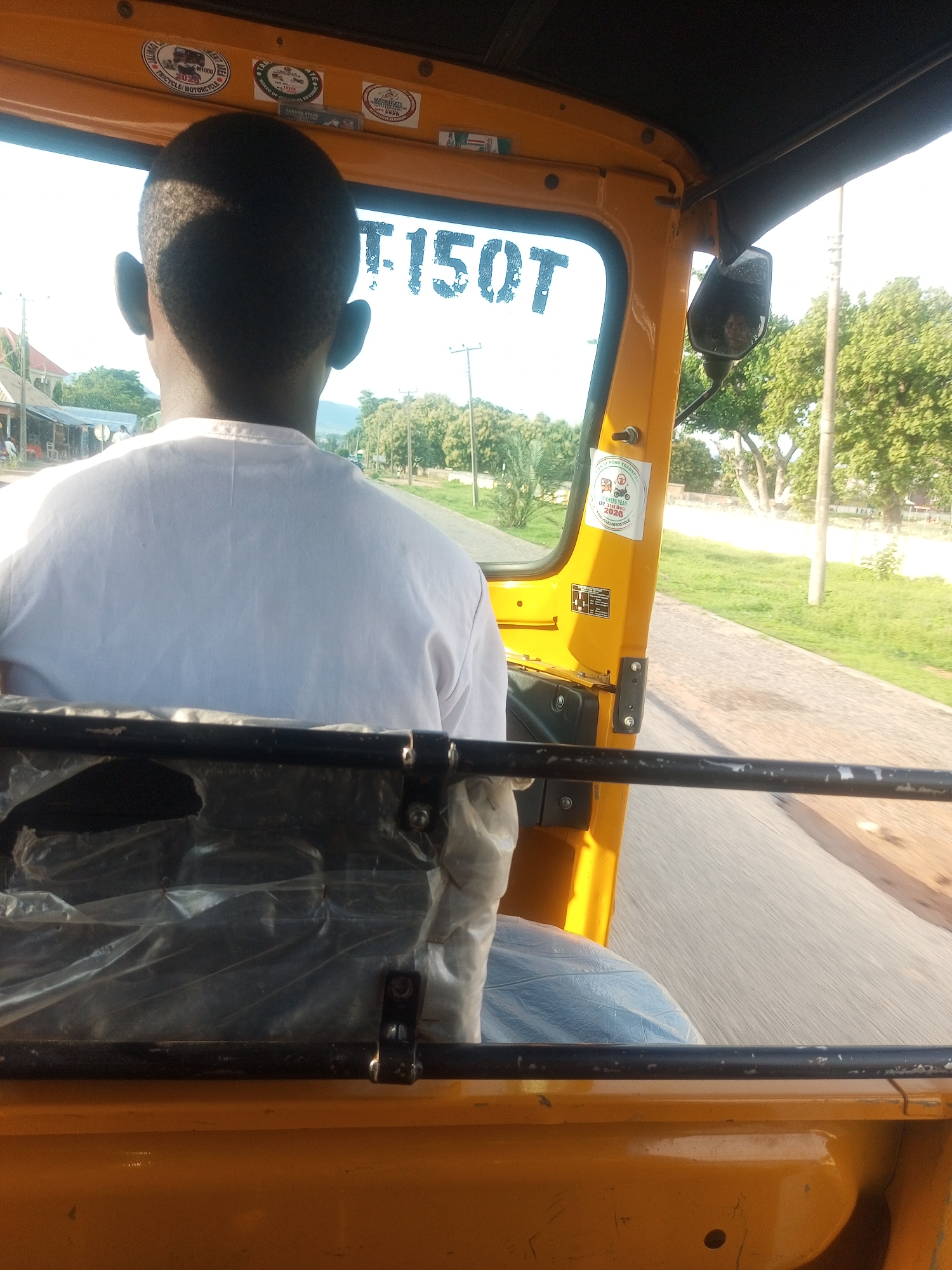 The image shows a person from behind, wearing a white t-shirt, seated in the passenger area of a yellow auto-rickshaw (also known as a Keke Napep). The auto-rickshaw appears to be in motion on a paved road. The scene is outdoors, with lush green trees and vegetation visible on the sides of the road. In the background, there are some buildings, suggesting a semi-urban or rural setting. The sky is bright, indicating daytime, and the sunlight casts shadows, suggesting a clear day.
The driver's head and shoulders are visible in the foreground through the windshield. The auto-rickshaw's interior is visible, with some plastic sheeting covering a seat or cargo area. There are several stickers on the interior of the auto-rickshaw, including one that reads "BOARD OF MINT TRICYCLE/MOTORCYCLE" with the year "2026" visible, and another that says "TARABA STATE ROAD TRANSPORT". This indicates the vehicle is licensed in Taraba State, Nigeria, and likely operating in or around Jalingo, the state capital. The overall impression is of a common mode of transportation in Nigeria, captured during a journey on a sunny day.