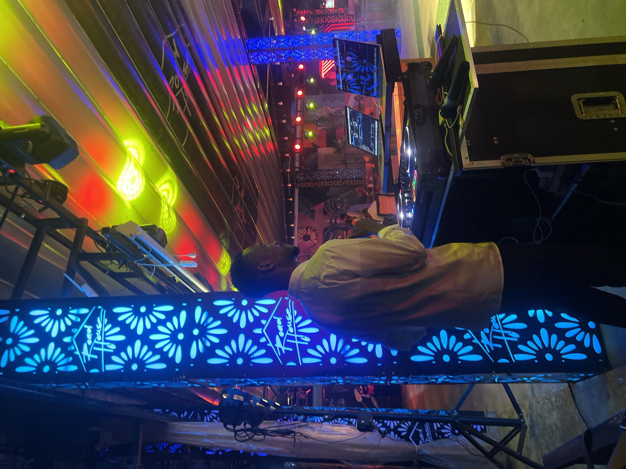 The image depicts a DJ set up in what appears to be a venue with vibrant lighting. A man, dressed in a light-colored shirt and dark pants, stands behind a DJ console. Two laptops are positioned on the console, along with a DJ mixer. Microphones are also visible on a shelf attached to the console. The background features a tall, illuminated column with a floral pattern and the logo "Road House". Colorful lights, a mix of red, yellow, and blue, illuminate the ceiling and parts of the environment, creating a lively atmosphere. The overall scene suggests an event or a performance taking place. The specific location in Howayi, Nigeria is not discernable from the image itself.