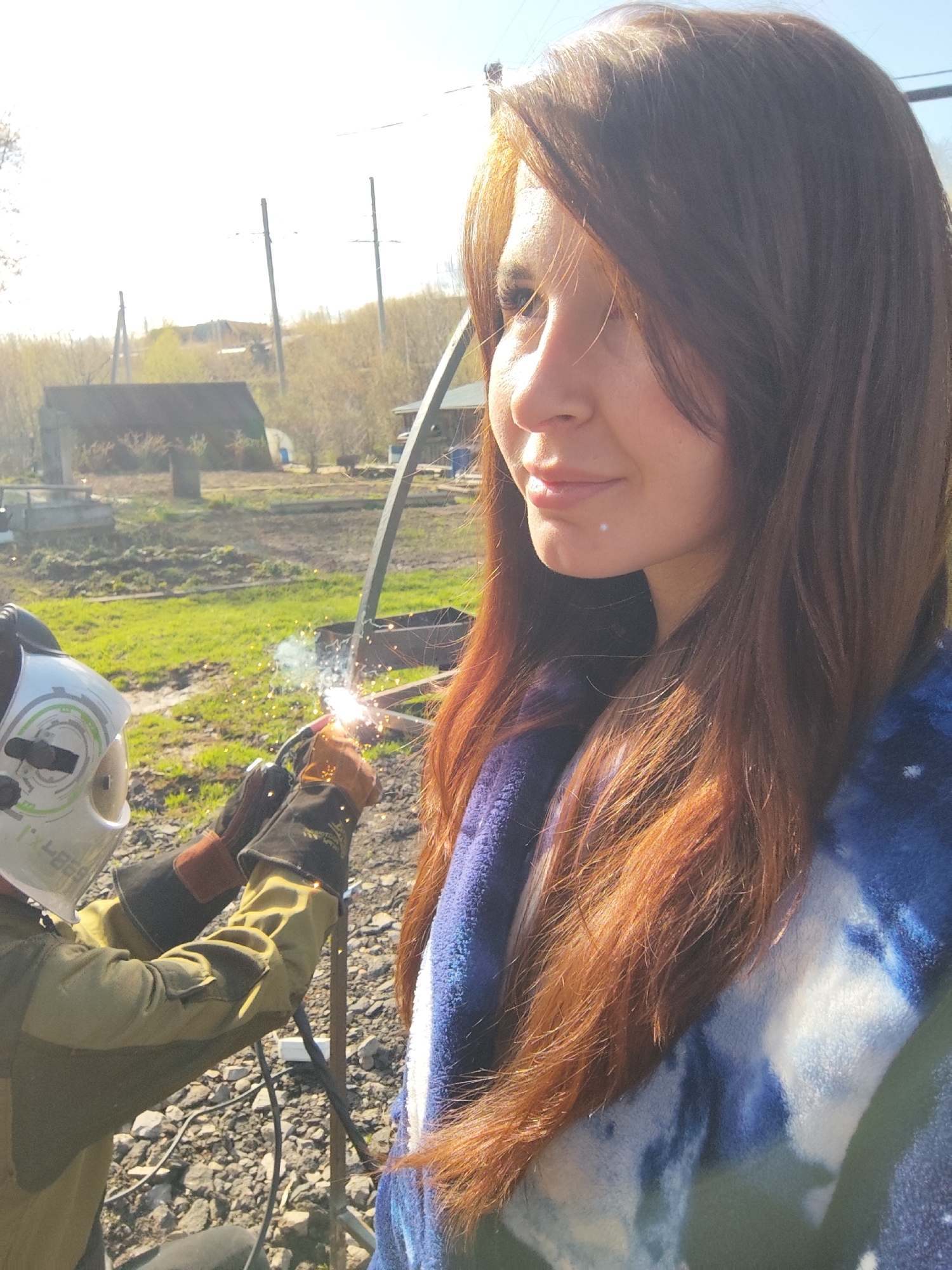 A medium shot captures a woman with long, auburn hair standing in what appears to be a residential or semi-rural setting, possibly located in Фоки, Russia, though this is based on the geographic information provided and not explicit features in the image itself. Her hair is styled with bangs and she is gazing to the left. She is wrapped in a blue and white patterned blanket.
In the background, slightly blurred, a person wearing a welding helmet and protective gloves is actively welding metal. Sparks are visible at the welding point. The person is wearing a light green jacket. The environment includes elements such as buildings, grass, trees, and utility poles in the distance. The ground in the immediate foreground is covered with gravel. The lighting suggests it is daytime. The woman is not interacting with the welder, suggesting she is a bystander or simply present in the scene.