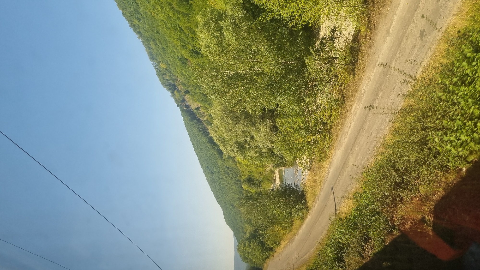 The image shows a road curving to the left in the foreground, bordered by lush green vegetation and dry grass. A shallow, rocky river flows alongside the road in the mid-ground, with a densely forested hill rising sharply on the right side of the frame. The sky is a clear, bright blue. The scene appears to be a rural or natural landscape, likely in or near Sasivka, Ukraine, given the context. The absence of any visible people or distinct man-made structures suggests a tranquil, natural setting.