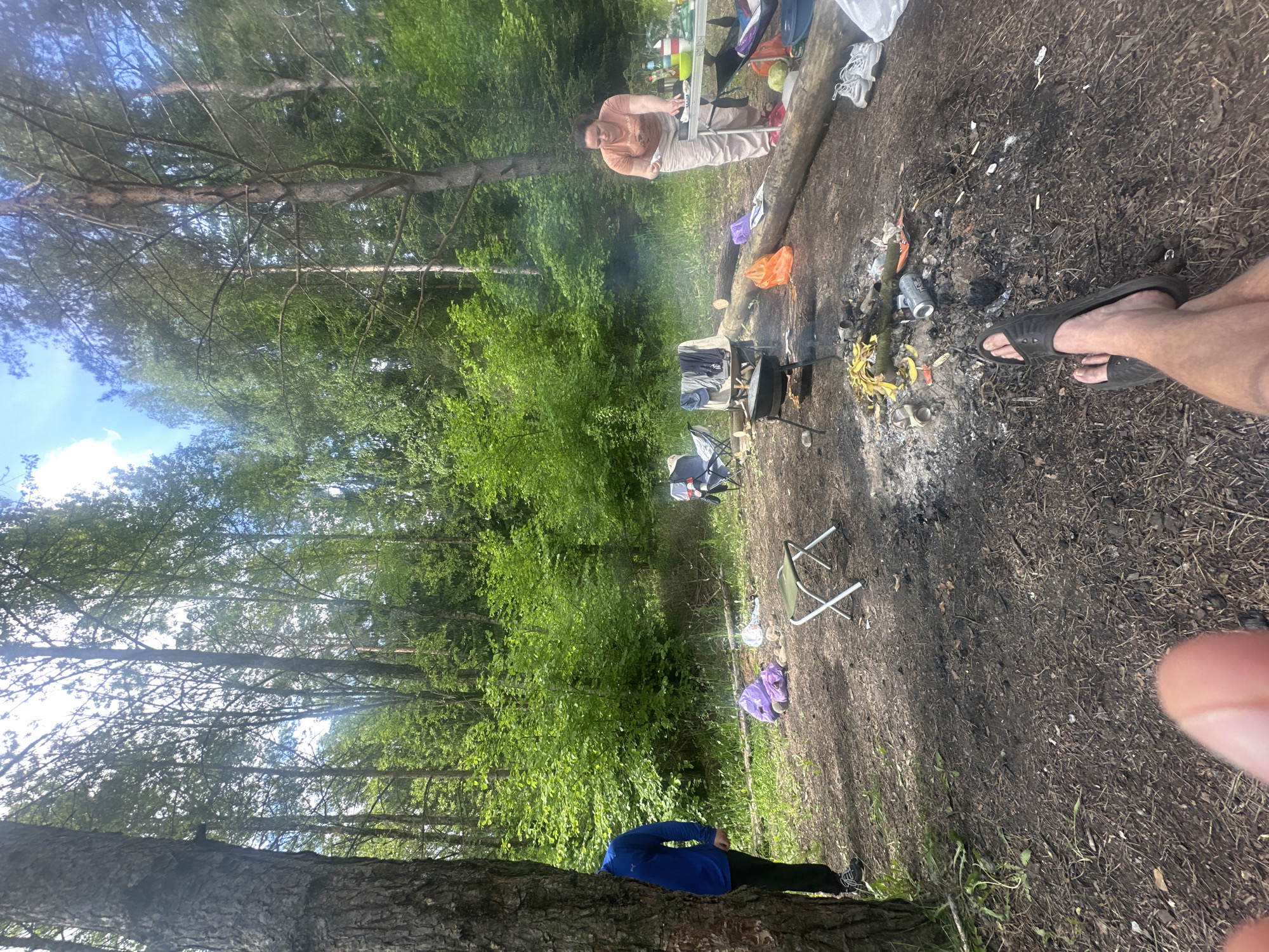 A campsite scene, possibly in Юзуфоўскі сельскі Савет, Belarus, shows a group of people gathered around a fire. One person in a peach-colored shirt stands near a table laden with supplies. A fire pit sits near the table, surrounded by scattered debris. A man in sandals is on the edge of the scene. Further into the woods, there are more people, chairs, and camping gear. The area is heavily wooded with tall trees. Another person is standing with their back to a tree wearing a blue jacket.