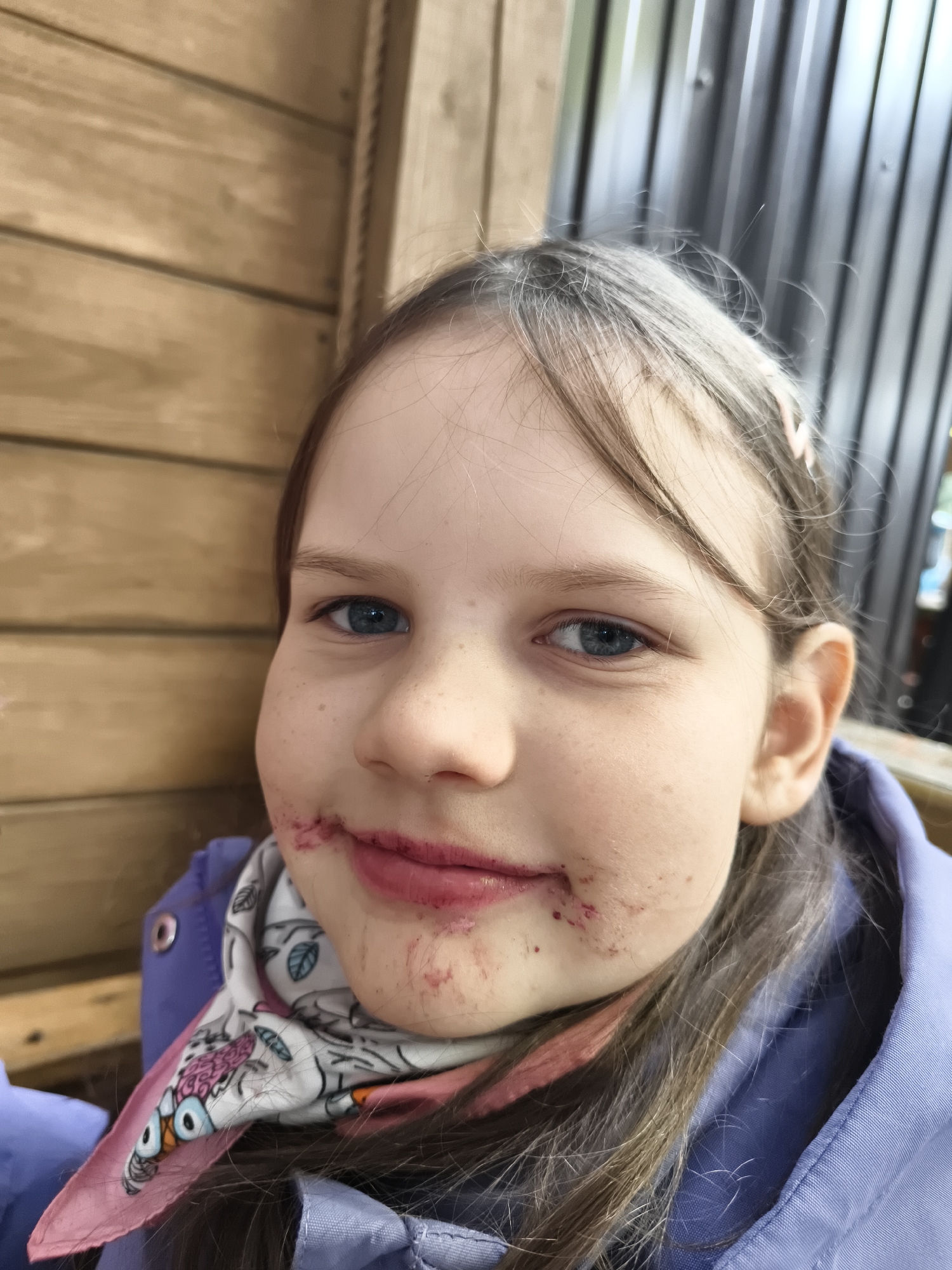 A close-up, eye-level portrait features a young girl with light brown hair and blue eyes, looking directly at the camera. Her face shows prominent red-purple stains around her lips and chin, indicative of having recently eaten berries or a similar food. She has a subtle, gentle smile on her face.
The girl is wearing a light purple or blue jacket, visible over her shoulders and chest. A white scarf with a colorful, possibly cartoonish, owl pattern and a pink border is tied around her neck. A light pink hair clip is partially visible in her hair on the right side of her head.
The background is divided into two parts: a light brown wooden wall with horizontal planks on the left, and a darker, corrugated metal surface on the right. The lighting suggests it is daytime. No text is visible in the image.