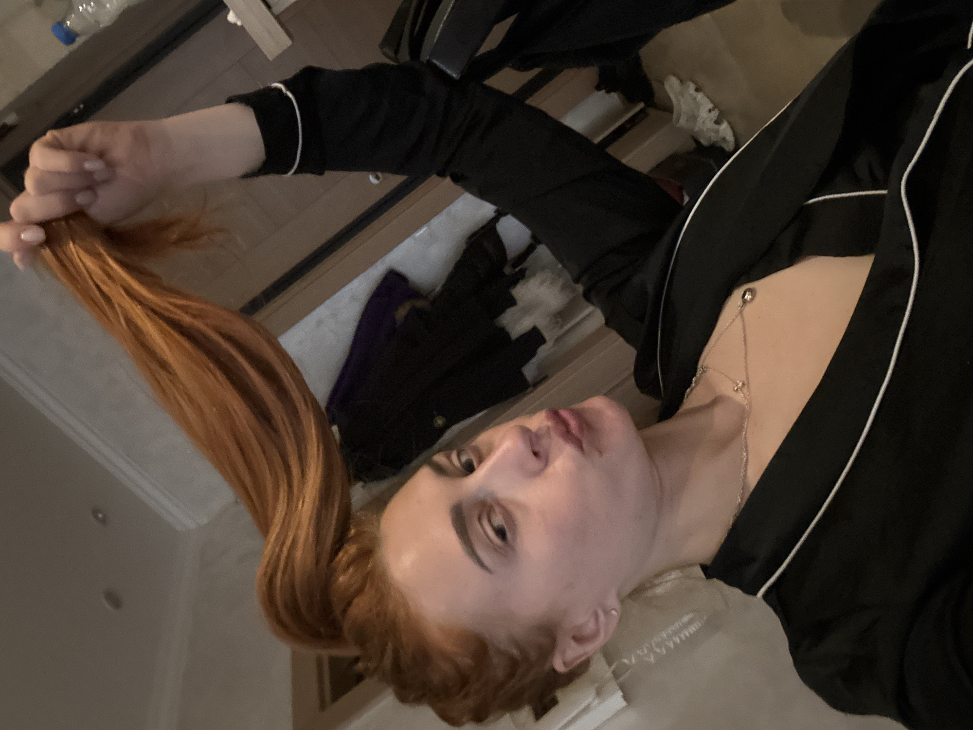An indoor, slightly high-angle medium shot features a female individual with long, reddish-auburn hair. She is seen from the chest up, with her left hand raised, grasping a portion of her hair and holding it vertically above her head. She wears a black top with white piping, resembling loungewear, and a delicate silver-colored necklace with a small pendant. Her expression includes pursed lips and a direct gaze towards the camera. A small stud earring is visible in her left ear.
The background shows light-colored walls and a ceiling with two visible recessed light fixtures. A wooden floor is partially visible in the lower left and right. To the right, a wooden surface supports a clear plastic water bottle with a blue cap. Other indistinct objects, possibly folded clothing or bedding, are visible behind her to the left, including some purple fabric. A dark object and a door frame are further in the background. White lacy fabric is also discernible on the floor in the lower right quadrant. The scene is located in Budva, Montenegro.