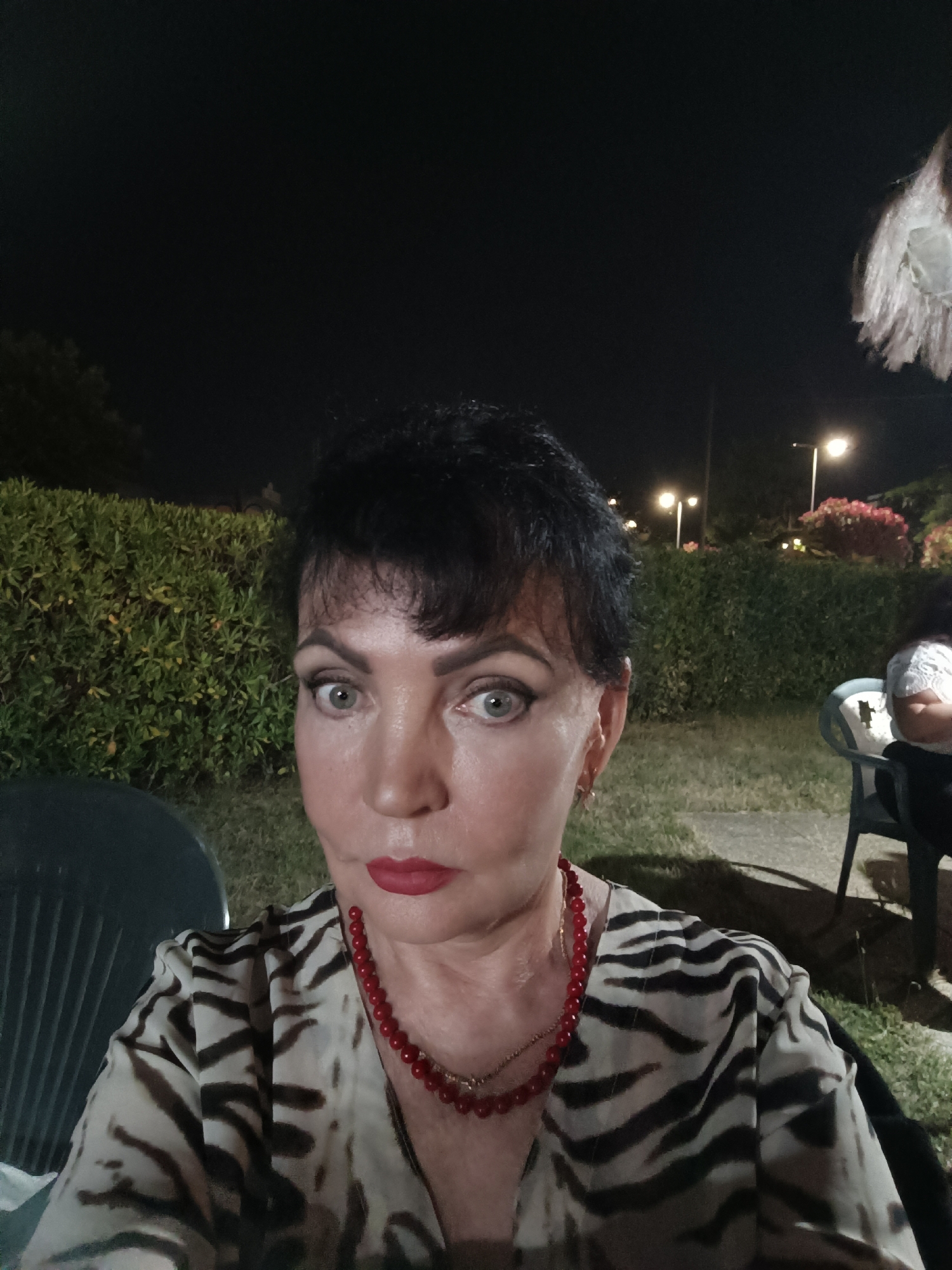 In the city of Giulianova, Italy, an adult female with short, dark hair, dark eyebrows, and prominent eye makeup is pictured from the chest up, at night. She is wearing a light-colored shirt with a black zebra-like stripe pattern and a red beaded necklace. Her lips are painted red, and she is looking directly at the camera with a neutral expression. Behind her, a dark green hedge is visible, and further in the background, there are streetlights and blooming pink flowers. To her right, a person is seated in a dark chair, with their back to the camera, wearing a white top. The scene appears to be outdoors, possibly a garden or patio area.