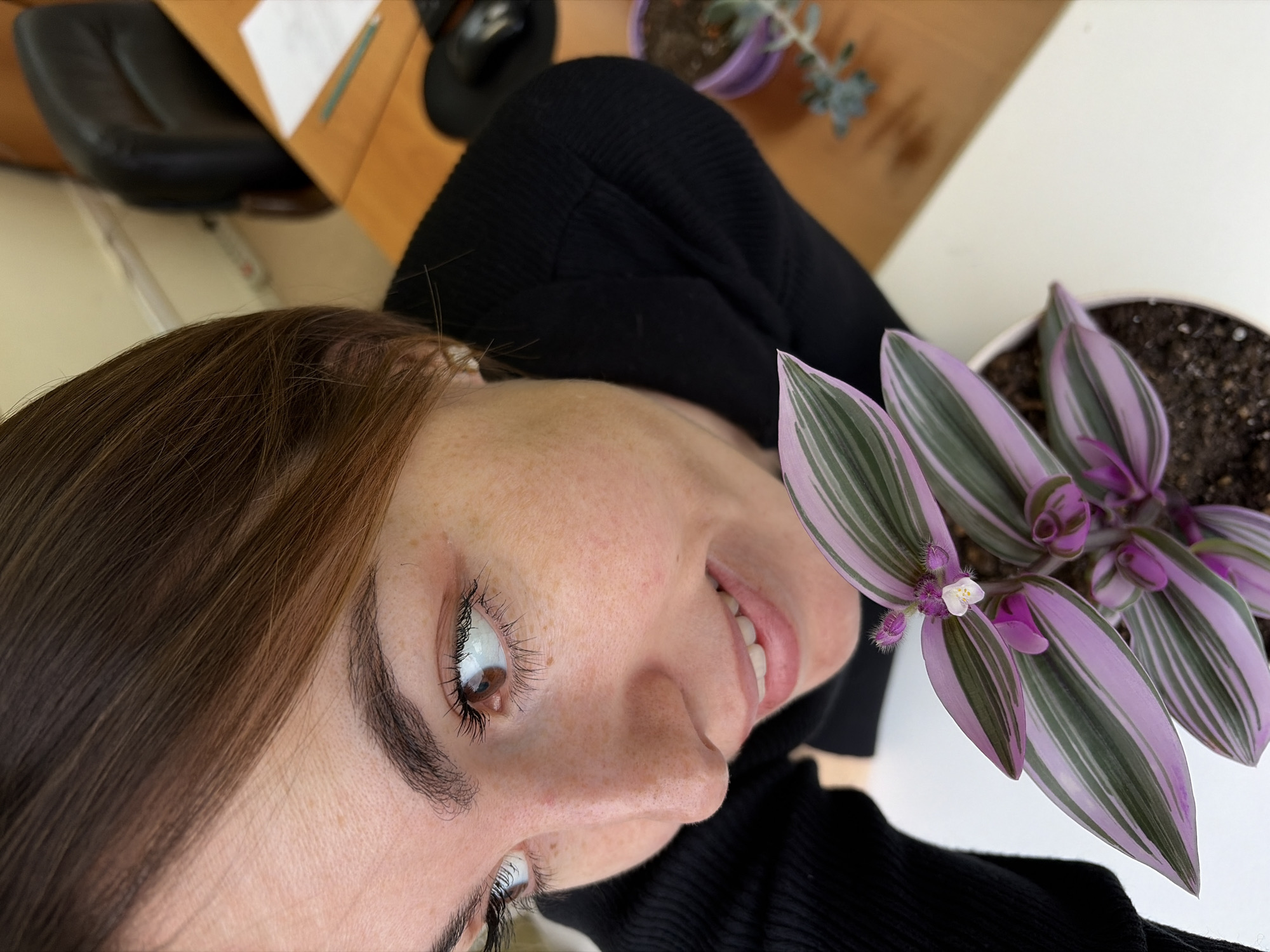 A close-up, high-angle shot focuses on a fair-skinned woman with freckles and brown hair. She is smiling slightly, and her brown eyes are visible. She is wearing a black turtleneck sweater. Next to her, on the right side of the frame, is a potted plant with purple and green striped leaves and small white flowers. The background includes a desk with a black computer mouse and a chair, as well as another potted plant, but those are out of focus. There is no information available to determine the location in the city of Izhevsk, Russia.