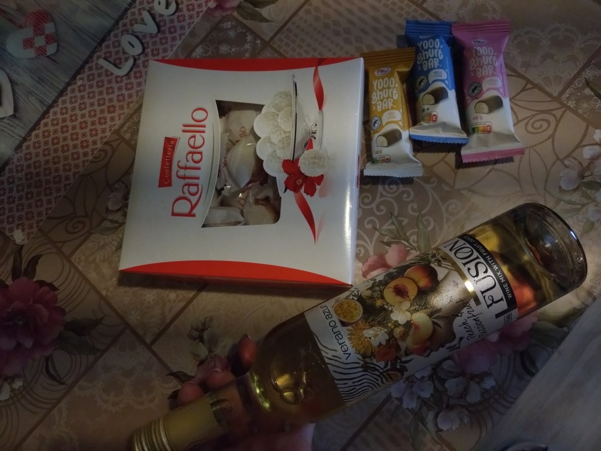 The image shows a flat lay of various sweets and a bottle of wine on a patterned tablecloth. In the center is a white and red box of RaffaELLO chocolates, with a clear window revealing several white, coconut-covered candies. To the right of the RaffaELLO box are three candy bars, one yellow, one blue, and one pink, all with the branding "YOOO. SHURT BAR." A bottle of "FUSION WINE MIX WITH FRUIT JUICE" featuring peach and passion fruit is positioned diagonally across the bottom right of the frame, held by a hand which is partially visible. The background tablecloth has a floral and geometric pattern. The scene appears to be indoors, likely on a table. There is no information in the image to suggest a specific location in Stara Zagora, Bulgaria.