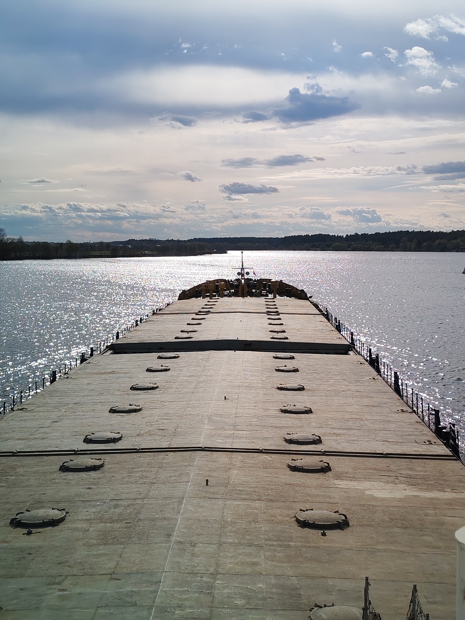 A high-angle shot presents the deck of a large vessel, possibly a barge or ship, extending towards the horizon. The deck is a vast, flat expanse, marked by a series of evenly spaced, circular hatches. Parallel rows of these hatches guide the eye towards the distant shoreline. The deck's surface displays a weathered texture, possibly indicative of rust or prolonged exposure to the elements.
The vessel is afloat on a body of water that reflects the sky's brightness, causing a shimmering effect. The shoreline in the distance is characterized by a dense tree line, suggesting a natural or forested area. Above, the sky is partially covered with clouds, some of which are illuminated by the sun. The overall lighting conditions indicate that the image was taken during daylight.
The location is not explicitly stated and remains unknown.