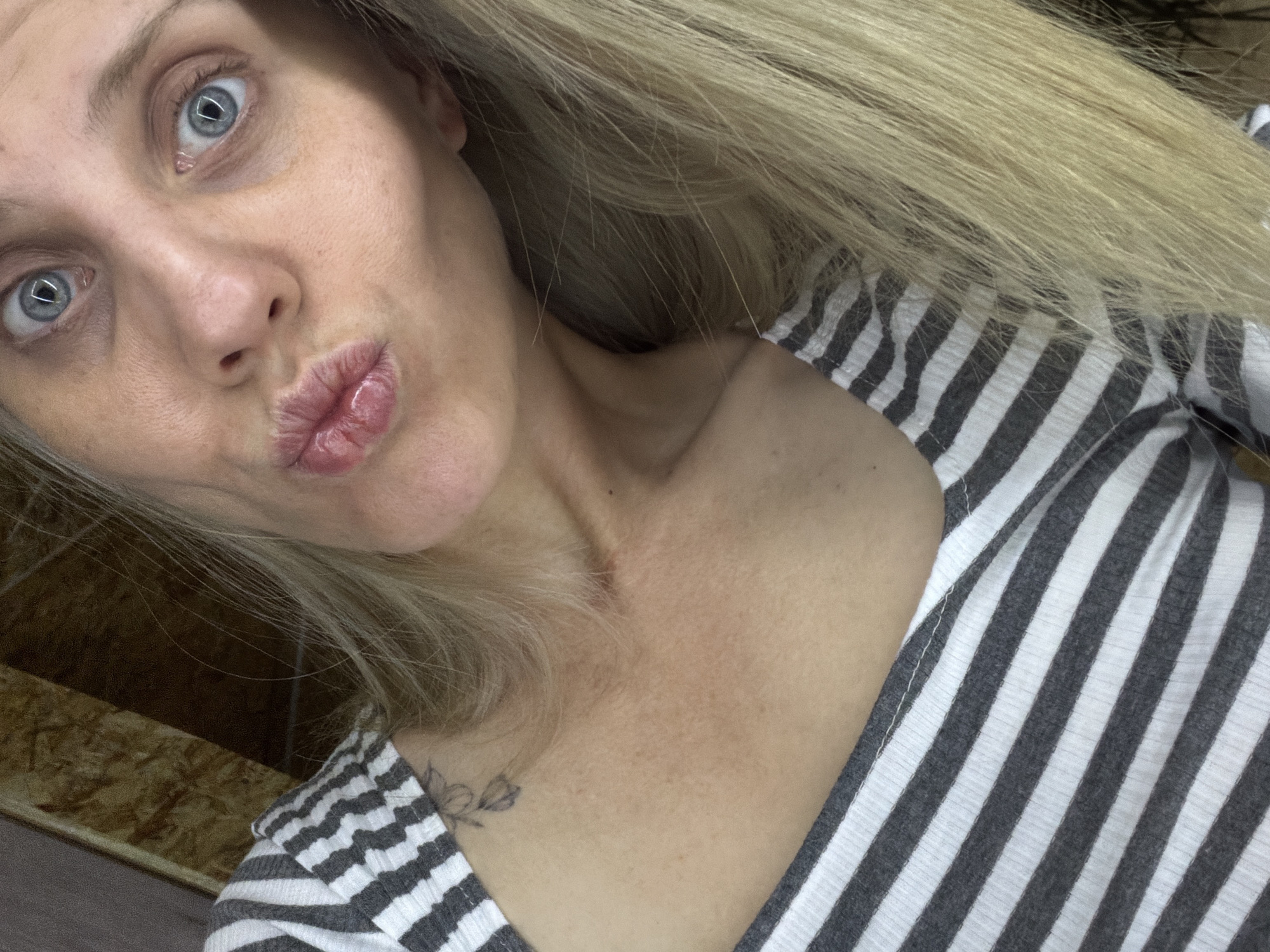 A fair-skinned woman with light blue eyes and blond hair is posing for a selfie. She is making a kissing face. She is wearing a striped grey and white shirt. There is a floral tattoo visible on her upper chest. The background appears to be a simple interior space with a wooden surface visible. The location of the city of Chaikovsky, Russia cannot be determined from the image.