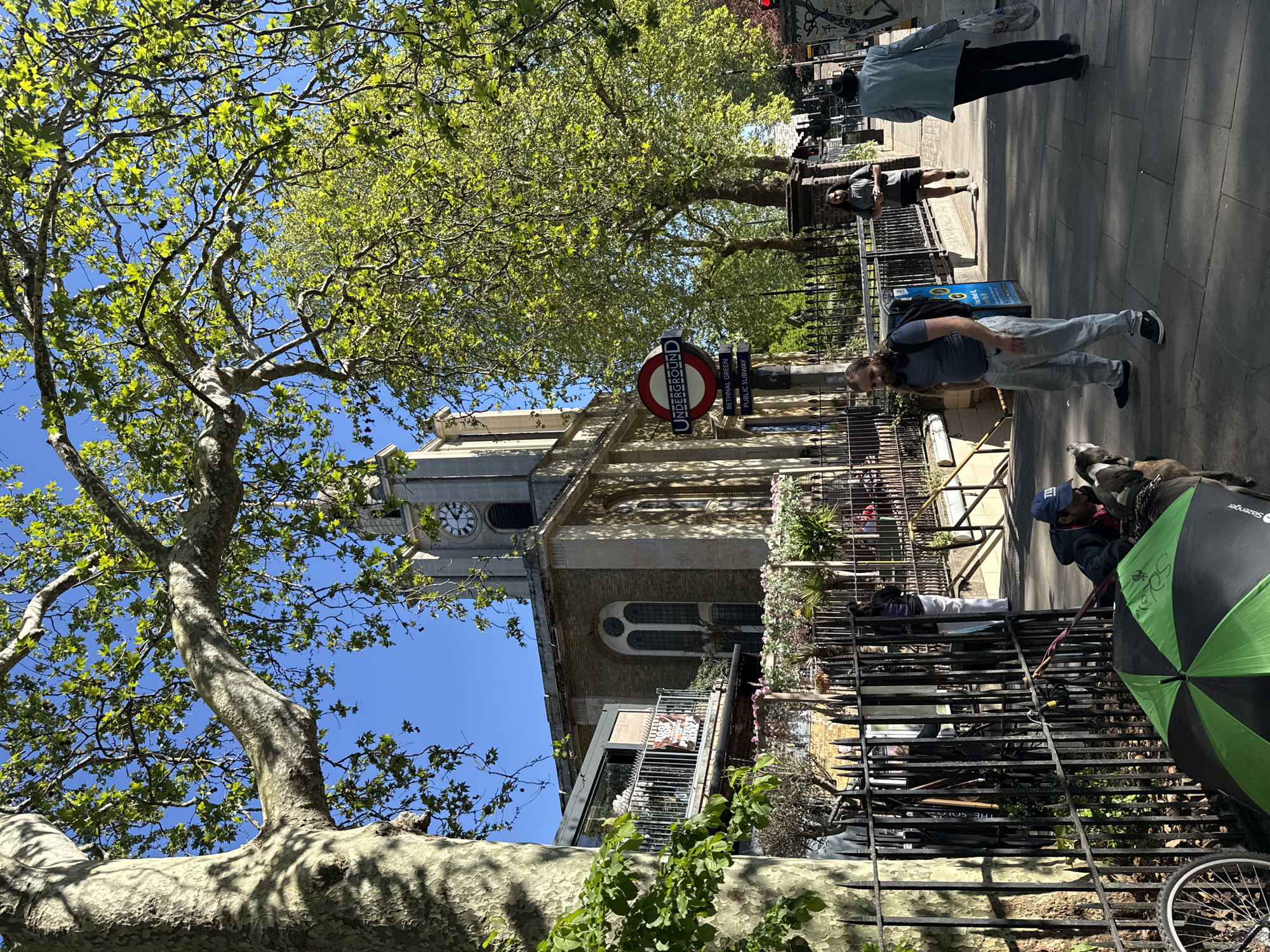 This image captures a vibrant street scene in Bethnal Green, London, on a bright, sunny day. A large, mature plane tree with lush green foliage dominates the foreground, its textured trunk and branches spreading across the frame against a clear blue sky.
In the mid-ground, a stone church with a prominent clock tower is visible, its clock indicating approximately 10:20. Below the church, a building features a balcony adorned with a sign that clearly reads "FREE ENTRY ALL DAY EVERY DAY". A black metal fence runs along the pavement, separating the street from the buildings.
Several people are present in the scene. A man with a beard and backpack, wearing a blue t-shirt and light trousers, walks along the pavement. Further back, another man in a grey t-shirt stands near the fence, and a person in a light jacket walks away. Closer to the viewer, a person in a blue cap is seen kneeling or sitting beside the fence, accompanied by at least two dogs, one of which is a large, dark breed. A black and green umbrella rests on the ground nearby, along with a bicycle wheel.
Key London identifiers are prominently featured: a classic red and blue London Underground roundel sign displays "Underground", and a signpost next to it points to "Bethnal Green" and "Public Subway". The strong shadows suggest a morning setting, characteristic of a pleasant day in the city.