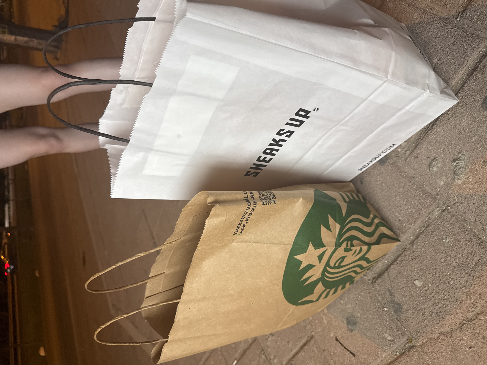 Two shopping bags are placed on a brick walkway at night. The bag on the left is made of brown paper and features the Starbucks logo. Text on the bag is partially visible and appears to be in Turkish, stating "STARBUCKS MOBİL UYGULAMASINI İNDİR, AYRICALIKLARINDAN FAYDALAN". The bag on the right is white and has the text "SNEAKS UP." printed on it in black, bold, sans-serif font. Below this, the URL "SNEAKS UP.COM" is visible. In the background, out of focus, a person's legs are visible, and a street with traffic lights can be seen. The location is identified as Kepez, Turkey.