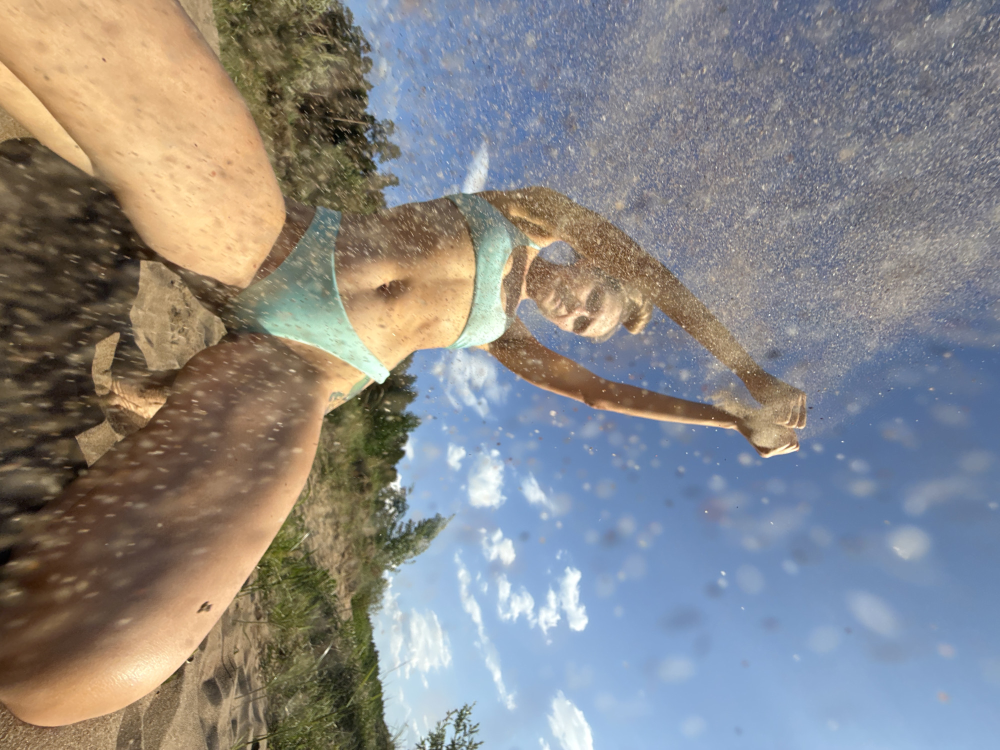 The photograph appears to be taken at an upward angle on a beach in Chaikovsky, Russia. A woman in a light blue bikini is the main subject. She seems to be either jumping or falling forward, with her arms extended upwards and outwards. Sand is suspended in the air around her, possibly kicked up by her movement. The background features a blue sky with scattered white clouds. Part of a grassy bank or hillside is visible to the left, suggesting a natural environment next to the beach. The woman's body is partially covered in sand, giving the impression of an action shot taken during a dynamic movement.
