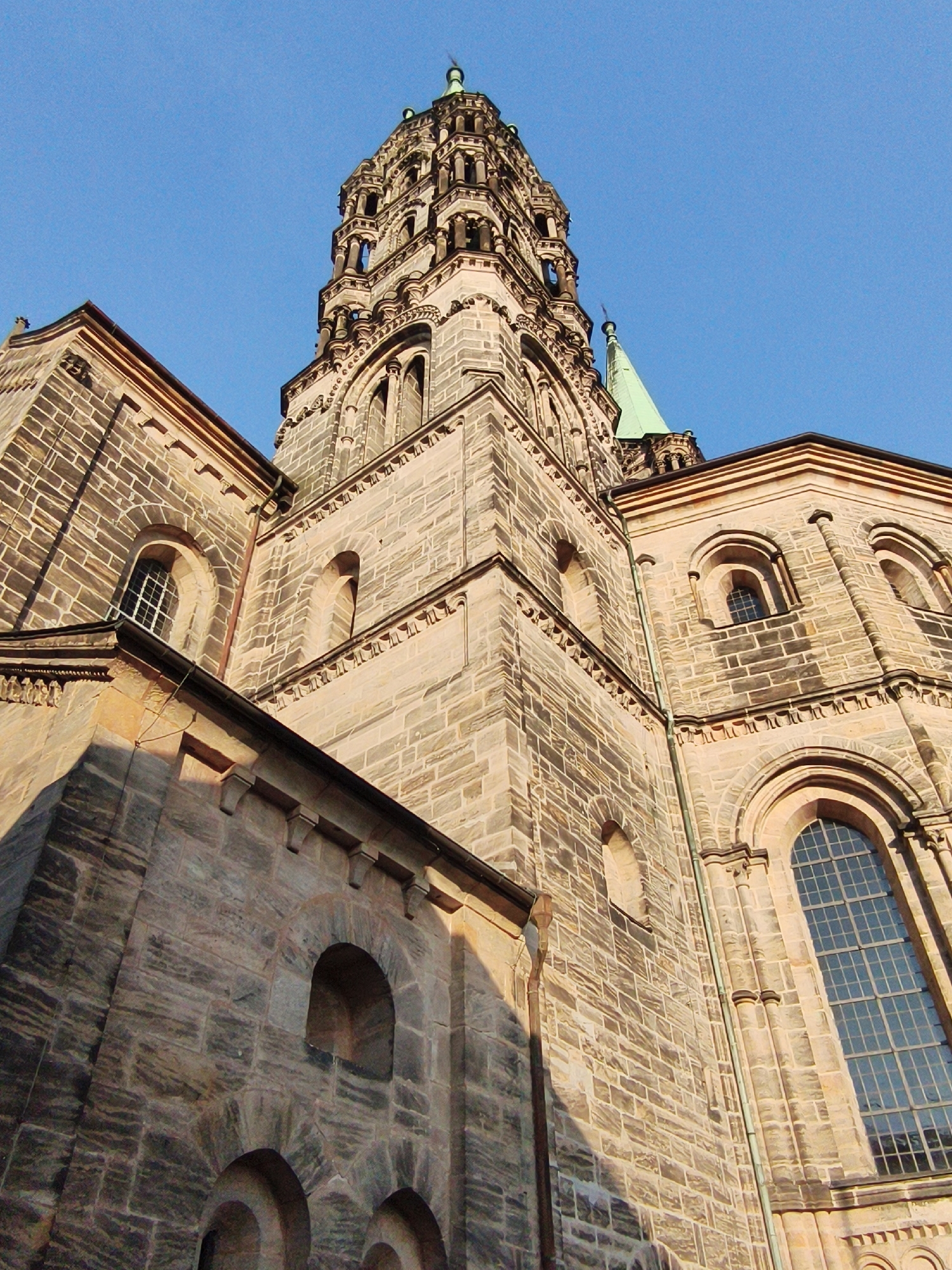 The image shows a low-angle, close-up view of the Bamberg Cathedral (Bamberger Dom), specifically its imposing tower and adjacent facade, against a clear blue sky. The architectural style is Romanesque, characterized by the heavy stone construction, rounded arches, and ornate detailing. The stone blocks are varied in color, ranging from light tan to darker gray, giving the structure a weathered and aged appearance. Sunlight casts strong shadows, highlighting the textures of the masonry and the intricate carvings around the windows and cornices. Several arched windows are visible, some with leaded panes, and one large, multi-paned window dominates the right side of the frame. The tip of a green-capped spire can be seen peeking above the main structure on the right. The perspective emphasizes the sheer scale and height of the cathedral.