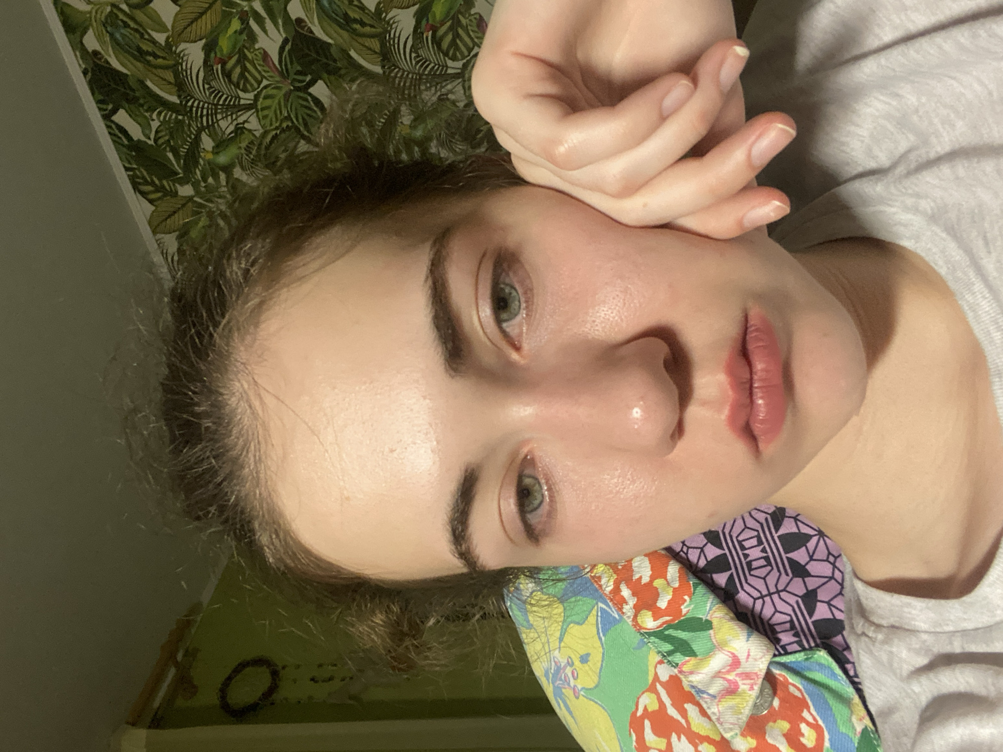 The media file shows a close-up, rotated, portrait-style photograph of a young person's face. The person has fair skin, light eyebrows, and green eyes. Their right hand is resting on their cheek and forehead, with their fingers splayed. They are wearing a light-colored t-shirt.
Behind the person, the background consists of patterned wallpaper with green leaves and birds, and a hint of a green wall or surface. A colorful, patterned fabric is visible in the foreground, partially obscuring the person's neck and shoulder. The lighting suggests an indoor setting, likely during the day or evening with artificial light. The overall mood appears calm and introspective.
No visible text is present in the image. The context provided places the location in Zavodoukovsk, Russia.