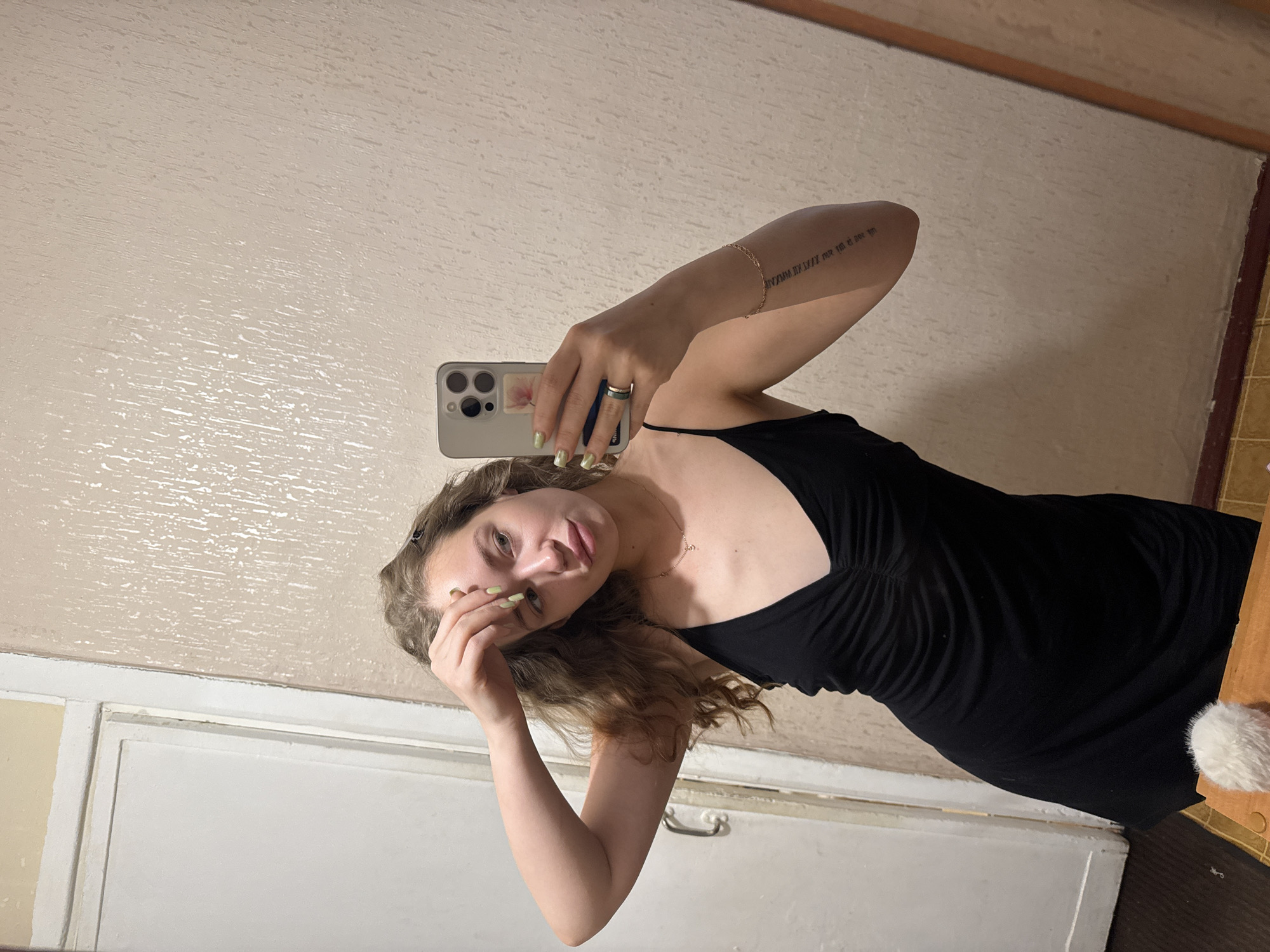 A young woman with wavy blonde hair is taking a selfie in a mirror. She is wearing a black spaghetti strap dress. She has light green nail polish on her fingernails and a delicate gold necklace. On her left arm, she has a tattoo in Roman numerals and a bracelet. She is touching her forehead with her right hand. The background appears to be an indoor setting, possibly a room in a residential building. The walls are textured and a pale beige color, and there is a white door visible on the left side of the image. The floor is a wood-like laminate. No specific location in the city of Chaikovsky, Russia can be determined from the image.