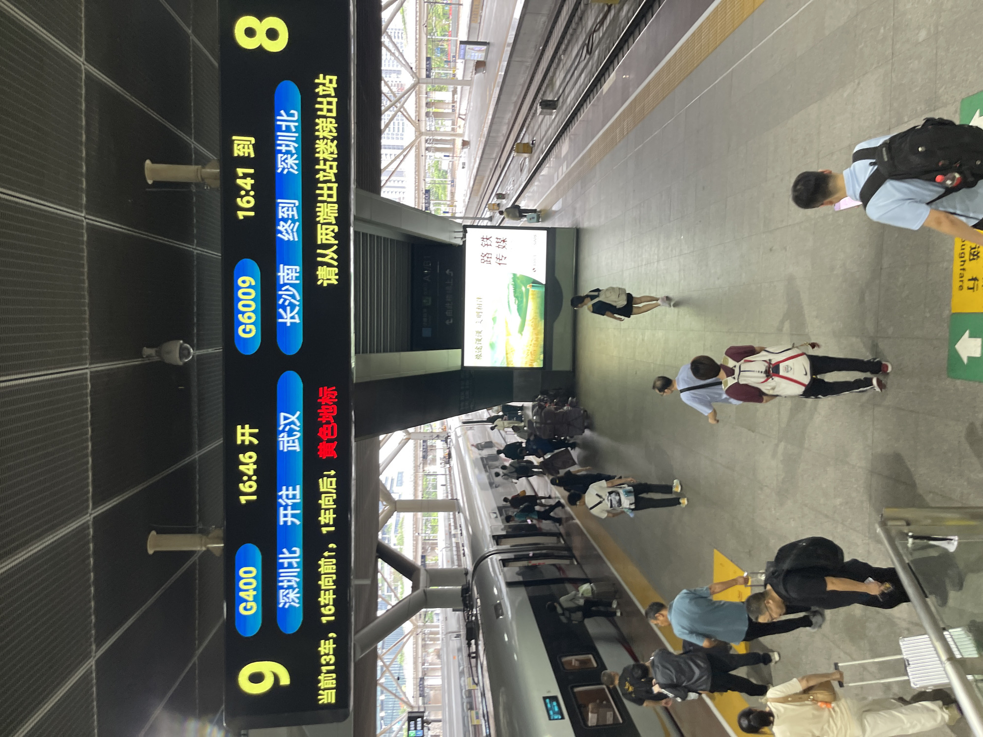 This image captures a bustling train station platform in China, likely during the daytime given the lighting. The primary focus is on a digital display board showing train information and the people waiting on the platform.
The digital display board prominently features train numbers like "G400" and "G6009," departure and arrival times ("16:46 开", "16:41 到"), and destinations ("深圳北 开往武汉", "长沙南 终到 深圳北"). It also includes directional information for passengers, such as "当前13车,16车向前,1车向后,黄色地标" and "请从两端出站楼梯出站". A train is visible on the tracks, suggesting an active station environment.
Several people are scattered across the platform, some with luggage, indicating travel. Their postures suggest waiting, moving, or possibly orienting themselves with the information provided. One person is looking at their phone, while others are walking or standing. The presence of luggage, backpacks, and shopping bags highlights the travel context.
A large advertisement panel with Chinese text, including "路铁 传媒" and "旅途漫漫之明相伴", is also visible, adding a layer of commercial or promotional content to the scene. The setting appears to be a modern, covered train station with clean gray flooring and visible ceiling structures. The overall atmosphere is one of transit and everyday activity at a busy transportation hub.