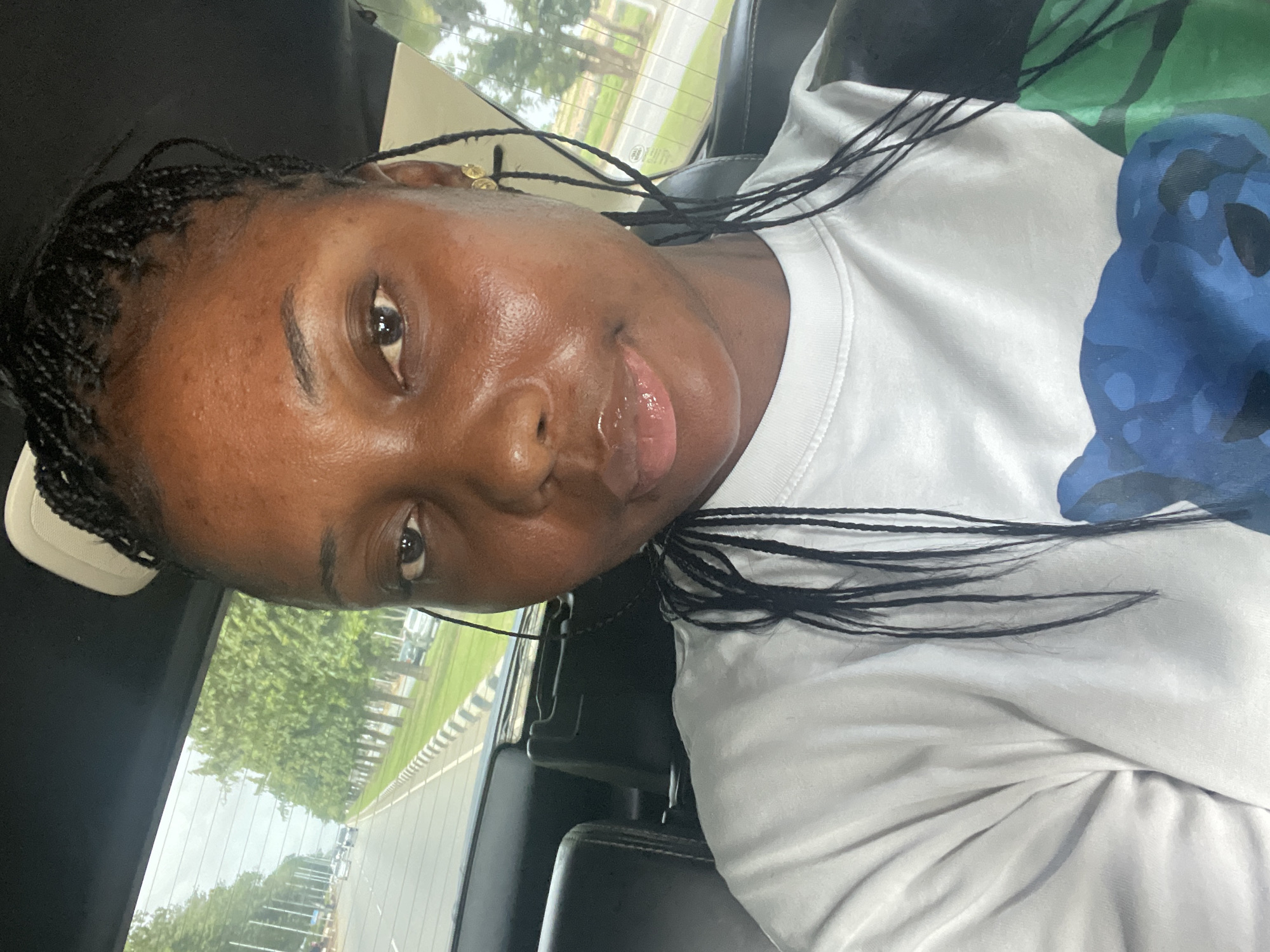 A young Black woman is depicted in a tilted, medium close-up shot from inside a vehicle, likely a car. She faces the camera directly, displaying a neutral to slightly serene expression with glossy lips. Her hair is styled in dark braids, some strands framing her face, and a gold-colored earring is visible on her right ear. She wears a white crew-neck t-shirt adorned with a partial blue and green graphic design on its right side.
The interior of the vehicle, characterized by dark upholstery, serves as the immediate setting. Through the rear window, a road scene unfolds, revealing white and black striped barriers running alongside the asphalt. Beyond the road, numerous green trees create a natural backdrop under a bright, possibly overcast, daytime sky. The overall lighting suggests it is daytime. Faint, unreadable characters or symbols are subtly reflected on the rear window glass. The image captures a candid moment inside a vehicle, consistent with being in transit or momentarily stopped, potentially within or near the Immigration Quarters in Nigeria.