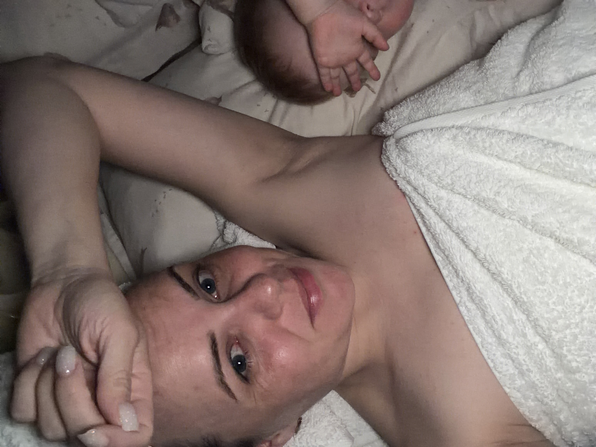 A woman lies on a bed covered in light-colored bedding and a white towel. Her head is tilted to the side, her hand resting on her forehead. A baby's head and hands are visible in the upper portion of the image. The image does not provide information regarding the location, specifically the city of Votkinsk, Russia.