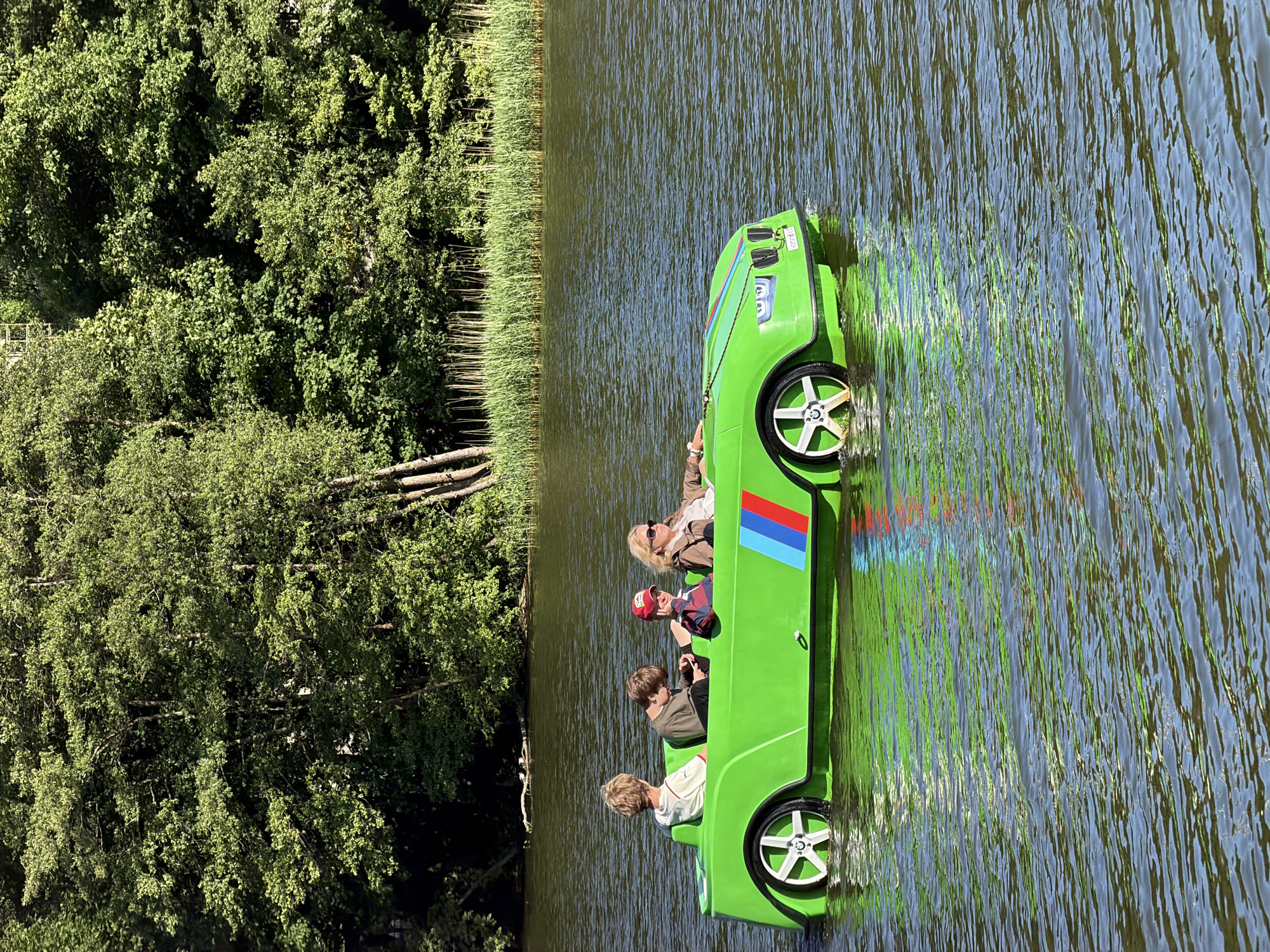 Four individuals, two adults and two children, are seated in a bright green, car-shaped pedal boat on a body of water. The boat features stylized headlights and a three-color stripe, reminiscent of BMW M colors, on its side. The water is rippled, and the background shows a dense line of trees and reeds. One child, wearing a red cap, is looking down. Another child is looking at a phone. An adult woman with sunglasses is steering, and a third person, possibly a child or young adult, is seated behind them, facing away from the camera. The scene appears to be a recreational activity on a sunny day. The location is identified as the city of Svetlogorsk, Russia.