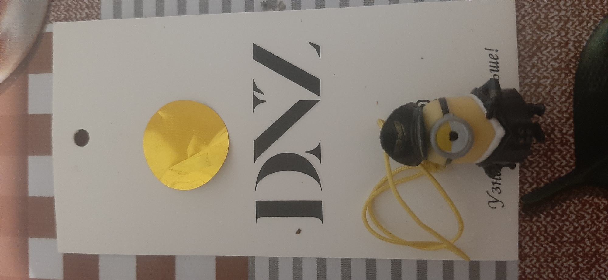 The image shows a white tag with the letters "DNZ" printed in large, dark grey font. Above the letters is a crumpled gold foil circle. Below the letters is a small Minion toy wearing a black hat and a suit, attached to a yellow string. Part of a Russian phrase, "Узна больше!", is visible below the Minion. The tag is placed on a surface with a checkered pattern. The image is taken from a top-down perspective. This image does not depict any identifiable location in the city of Yessentukskaya, Russia.