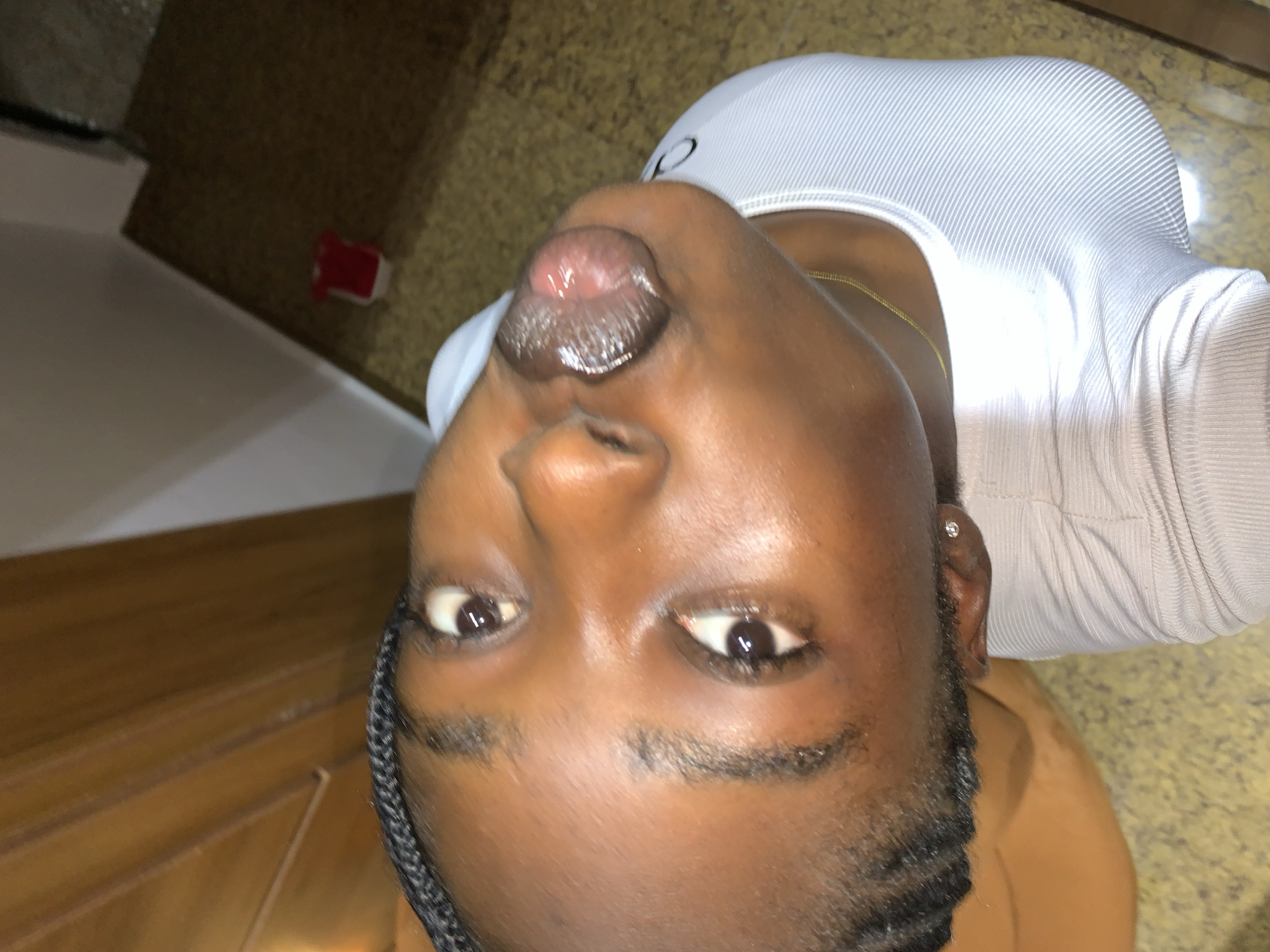 The image shows a close-up, high-angle view of a young Black person with braided hair, wearing a white ribbed top and a gold chain. The person's face is the primary focus, with their eyes open and looking upwards. Their lips are slightly parted, revealing their tongue. The background appears to be an indoor setting, with a wooden surface and a tiled floor visible. A small red object is on the floor in the distance. No specific details about the location in Gura Top, Nigeria, are discernible from the image.