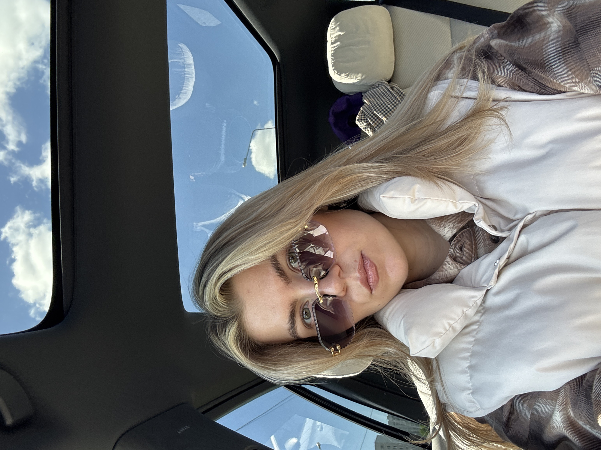 A woman with blonde hair is sitting in a car. She is wearing sunglasses, a white puffy jacket, and a plaid shirt. The car has a sunroof that shows a cloudy sky. There is a pillow and a hat on the backseat. The location of the photo is in the city of Unknown, Unknown.
