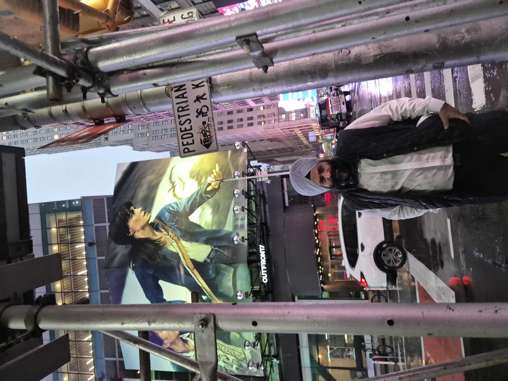 The image depicts a street-level view in Times Square, New York City, at dusk or during a rain shower, as evidenced by the wet pavement and reflective surfaces. In the foreground, a man wearing a grey turban, a white collared shirt, and a black quilted vest stands facing the camera. He has a dark beard and is looking directly at the viewer with a neutral expression. His left hand is tucked into his vest pocket.
Behind the man, several elements of the urban environment are visible. Large digital billboards dominate the background, featuring a prominent advertisement with a woman in a denim jacket. Below the billboards, a white SUV is parked on the wet street, with its headlights on. Further down the street, a red truck is visible, along with other vehicles and streetlights. A "PEDESTRIAN" sign with directional arrows is affixed to a pole in the middle ground, indicating pedestrian crossing areas. Scaffolding and utility poles are present in the foreground and midground, suggesting ongoing construction or infrastructure. The overall atmosphere is characterized by bright artificial lighting from the billboards and streetlights, contrasting with the dimming natural light.