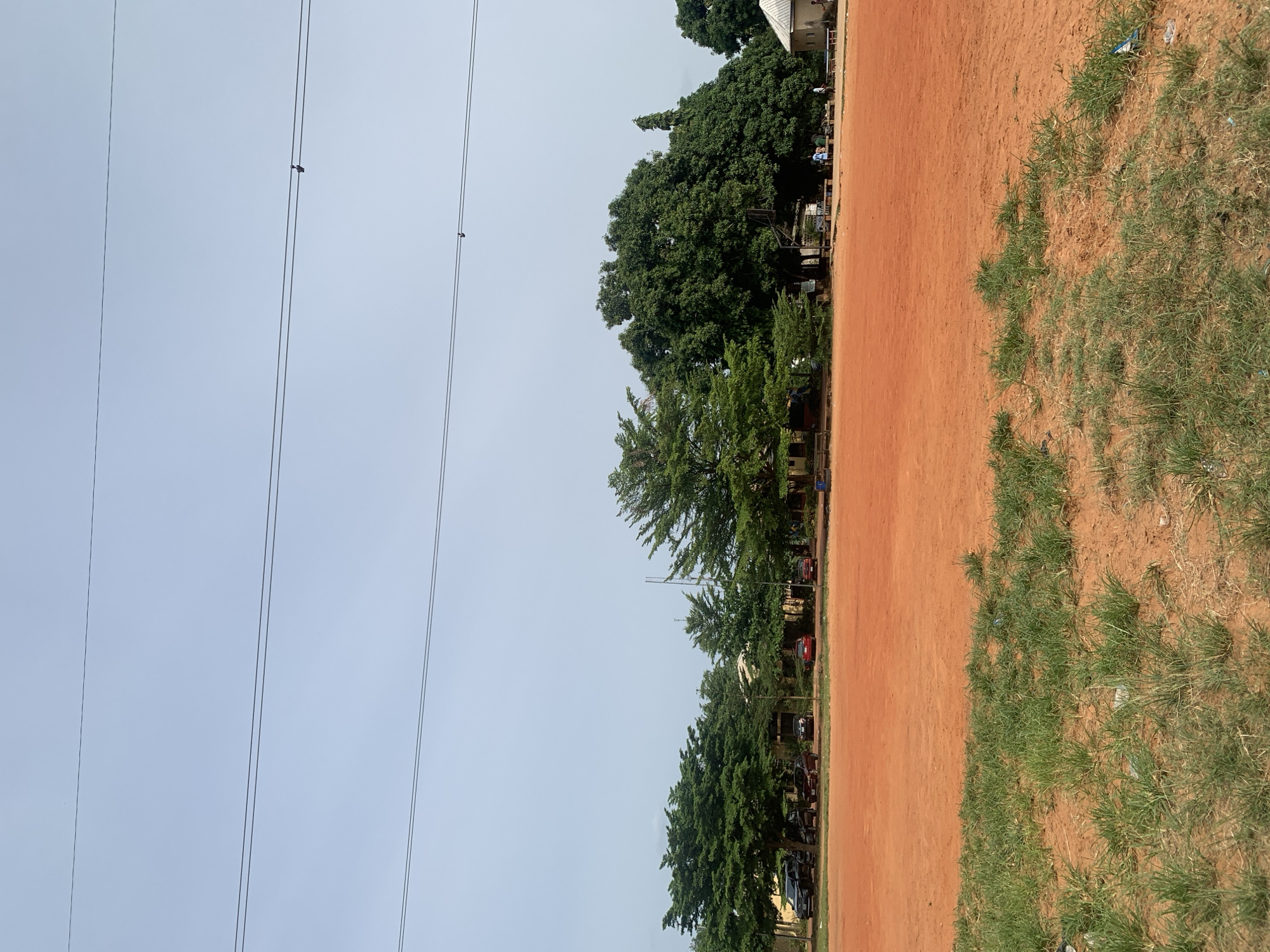 The image captures a wide shot of an outdoor area, likely a school or community ground, dominated by a large, flat expanse of orange-red dirt in the foreground. A sparse patch of green grass grows along the bottom edge, with some visible litter. In the mid-ground, a line of lush green trees forms a backdrop, with several buildings partially visible behind them. Some cars are parked in a line in front of the buildings, and a few individuals can be seen walking or standing near them. A basketball hoop stands beneath one of the larger trees. Power lines stretch horizontally across the pale, overcast sky, with two small objects hanging from the middle wire. The scene appears to be a typical day in an outdoor urban or semi-urban environment.